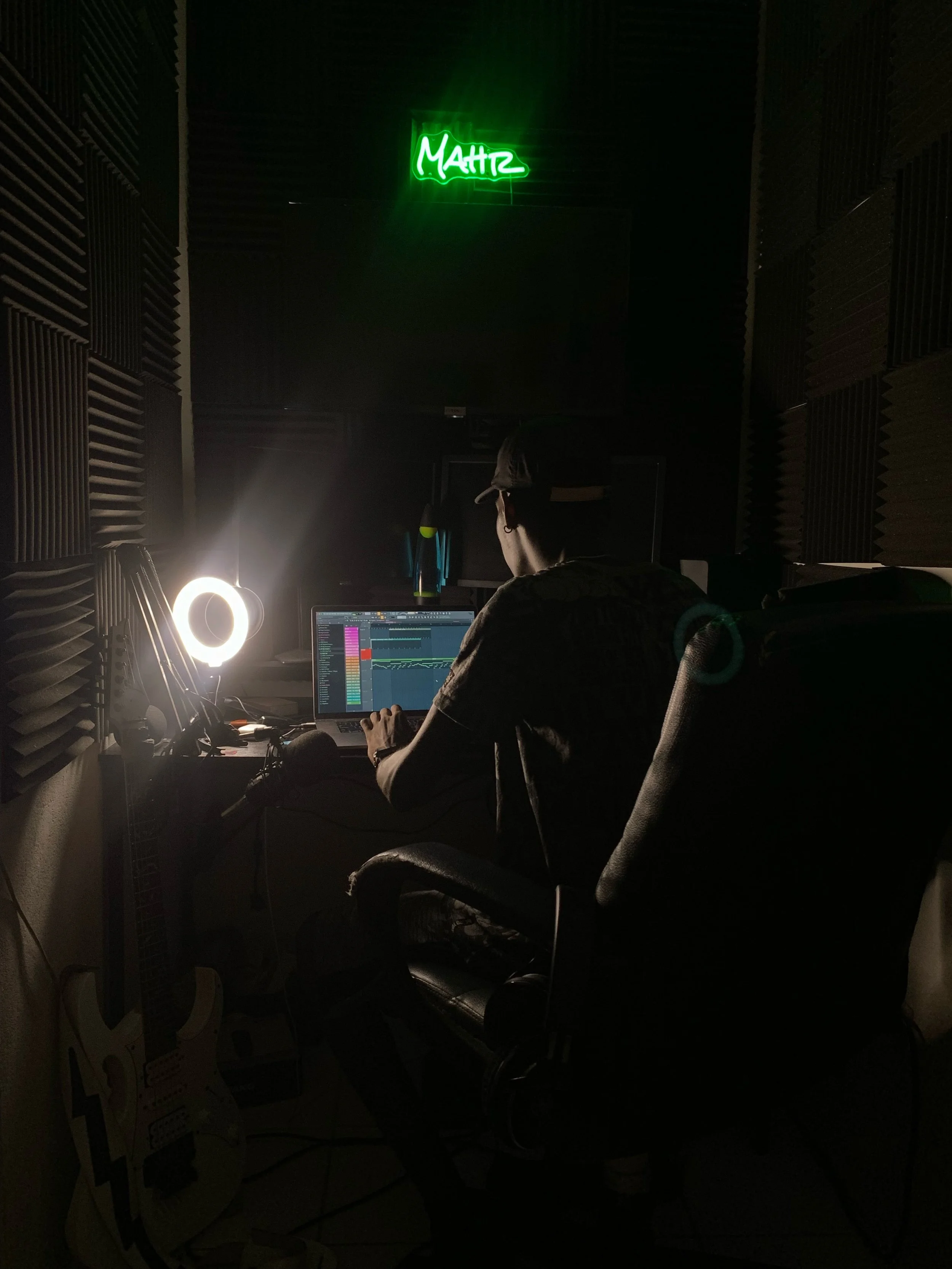 A man sitting in a dark music studio, working on a laptop with music production software, illuminated by a ring light. A guitar is on the floor, and a green neon sign spelling 'MATIZ' is on the wall.