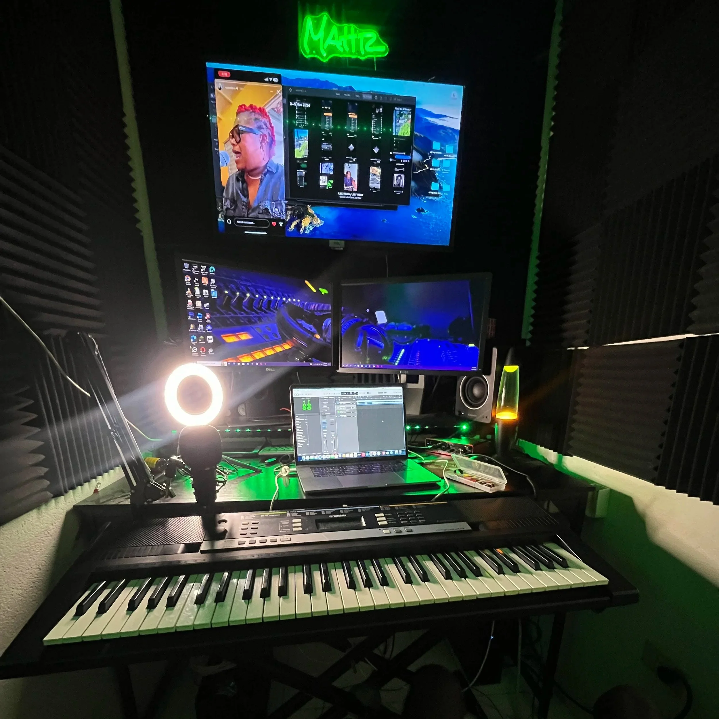 Music production studio with a keyboard, three monitors, a laptop, a microphone, studio speakers, a ring light, a lava lamp, and acoustic foam panels, illuminated by green LEDs and a neon sign that says 'MAHIZ'.