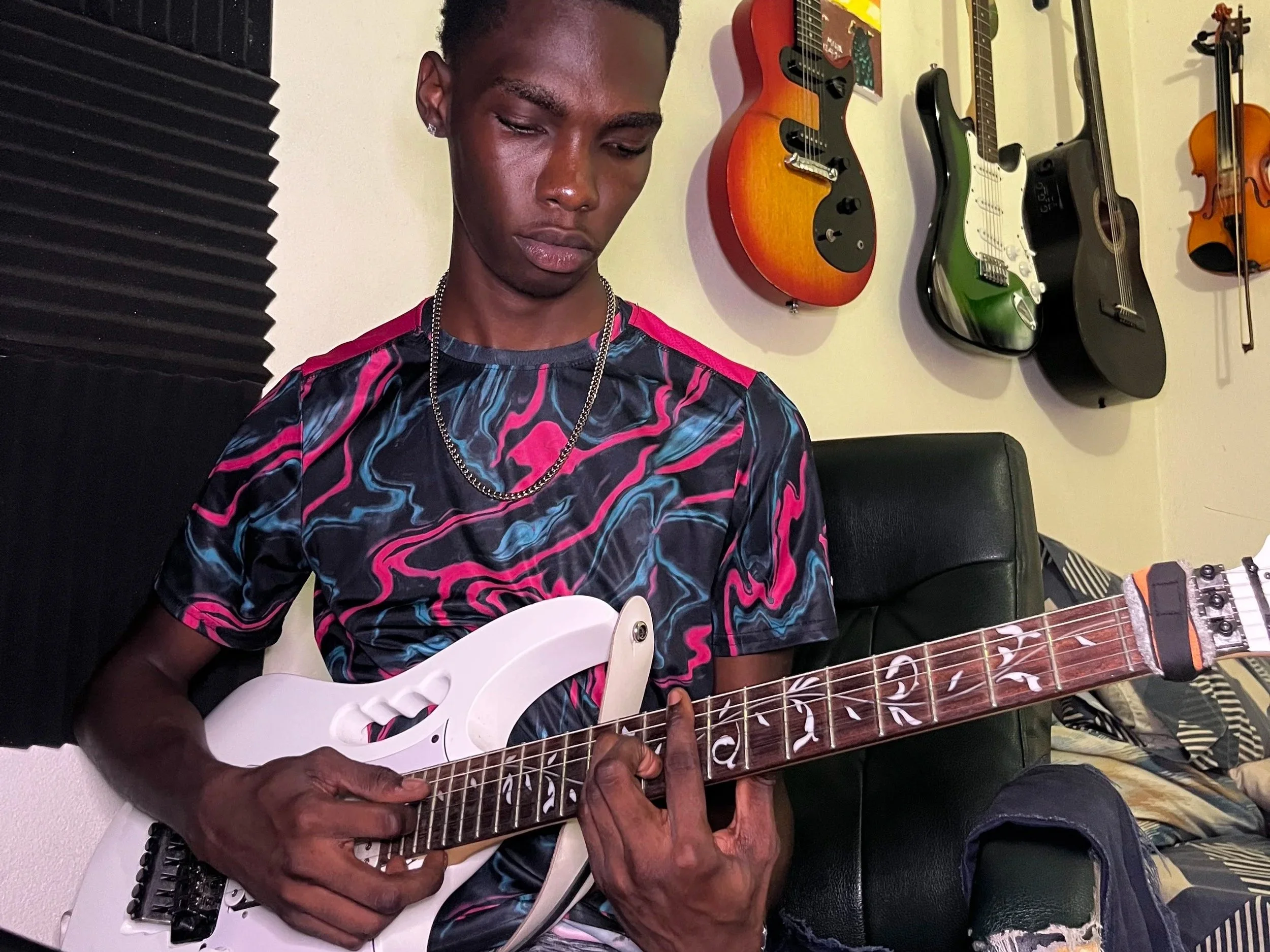 Young person sitting on a black chair, playing an electric guitar indoors, with various guitars hanging on the wall behind them.