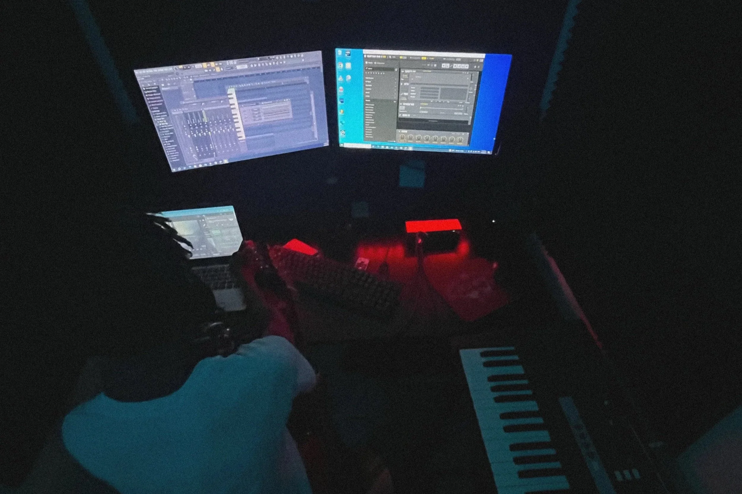 A person sitting at a desk with multiple computer screens displaying music production software, a keyboard, a laptop, and electronic music equipment, with dim lighting and red ambient lights.