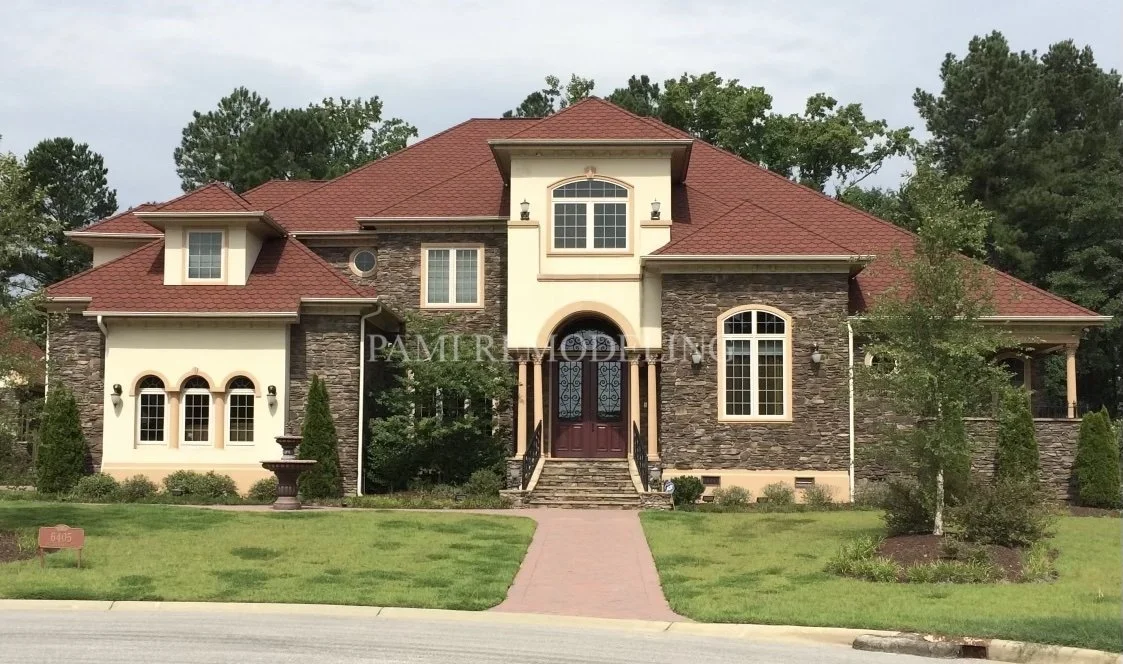 Large house with a red shingle roof, stone and stucco exterior, front steps leading to a double door, surrounded by trees and a well-maintained lawn.