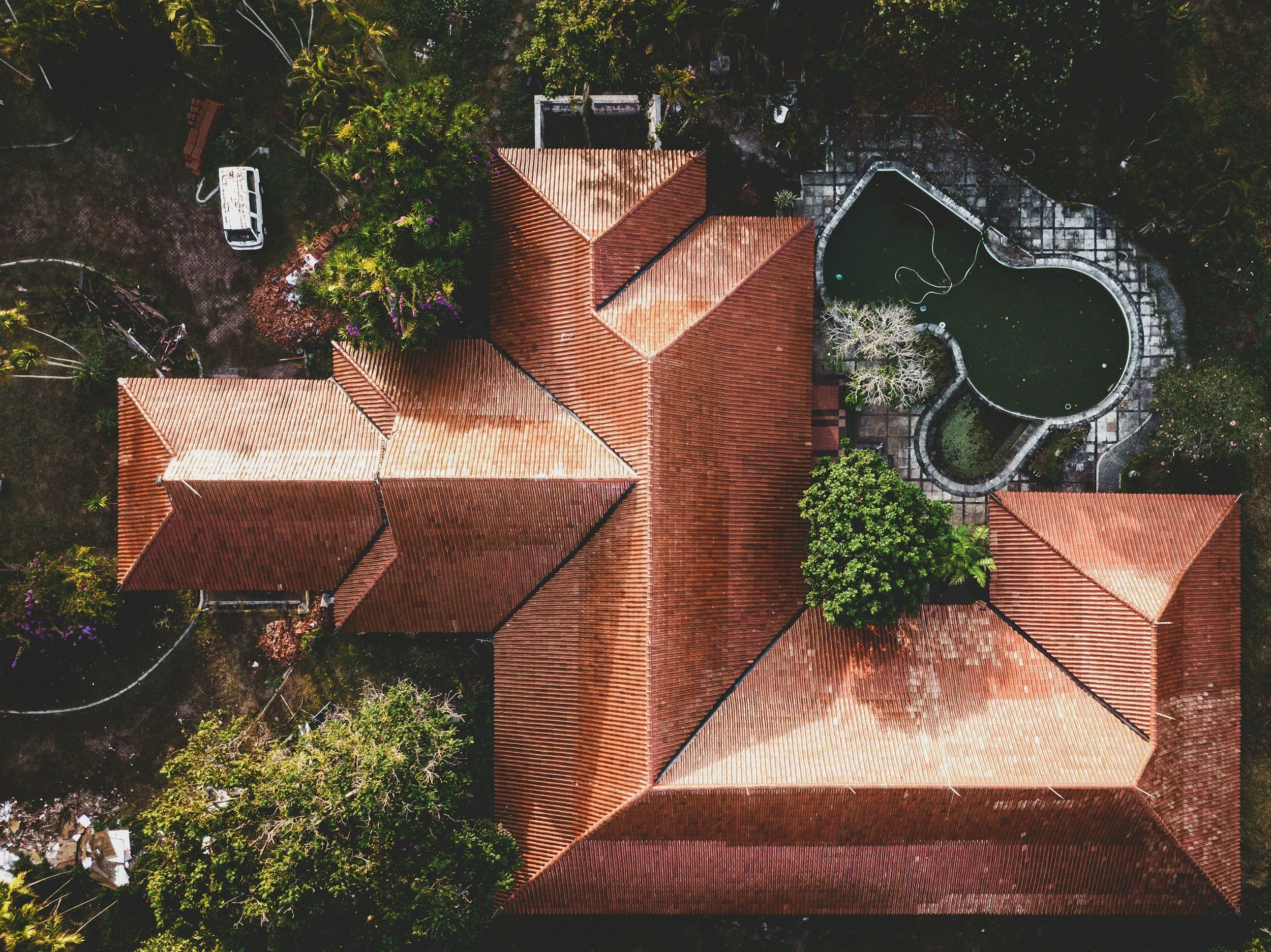 Aerial view of a large house with a terracotta tile roof, surrounded by a garden with trees, plants, and a swimming pool shaped like a kidney.