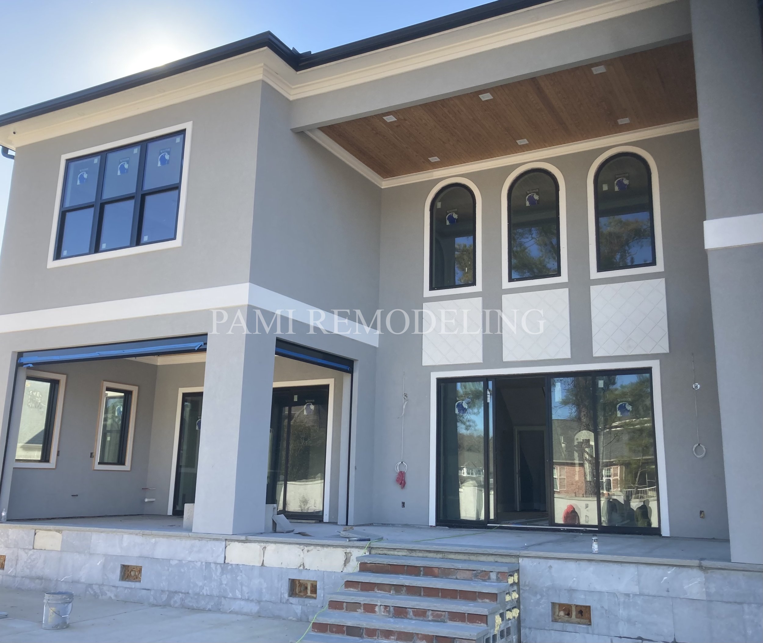 Front view of a two-story house under construction with gray walls, large sliding glass doors, and arched windows, featuring a covered porch area with a wooden ceiling and brick steps leading up to the entrance.