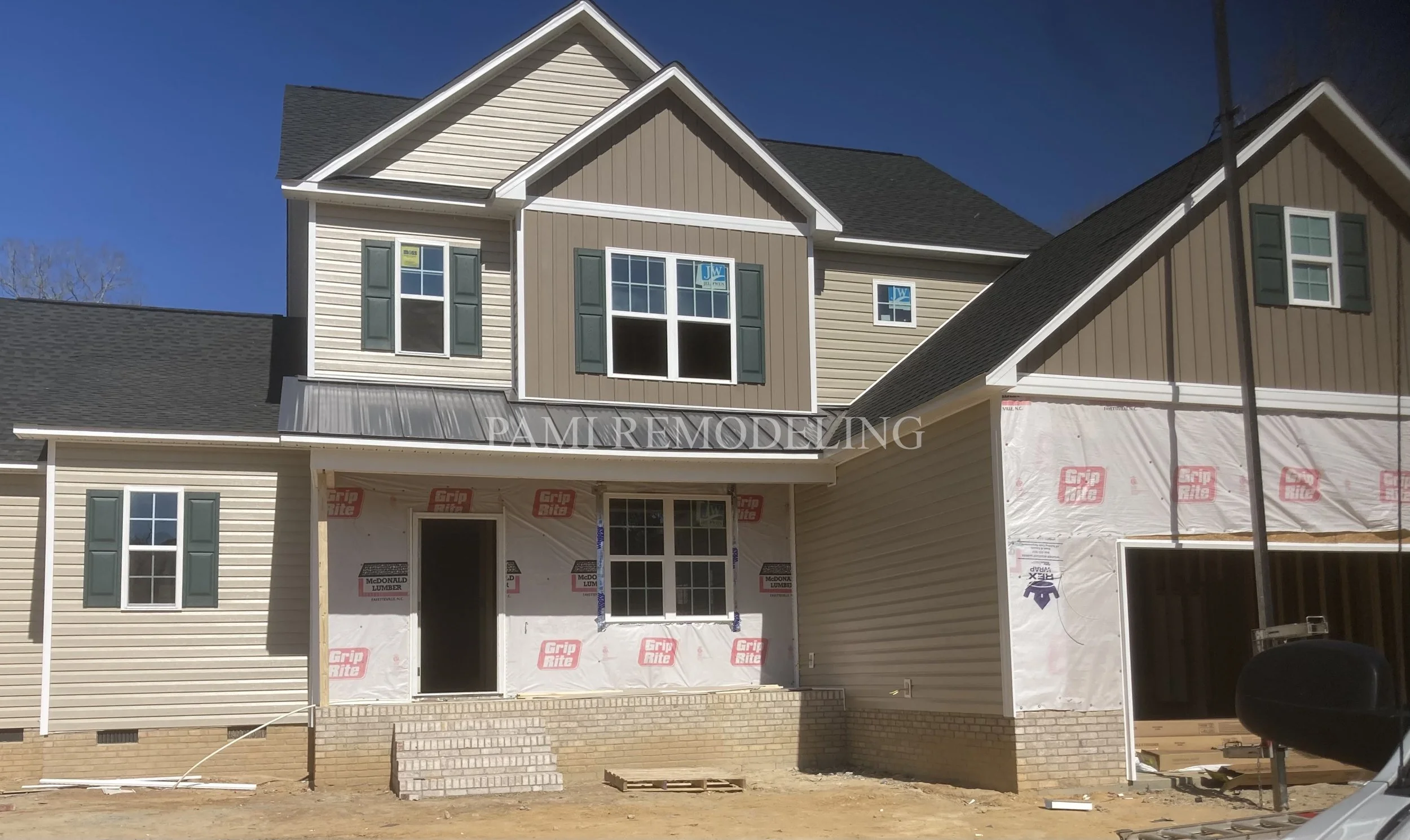 Under-construction multi-story house with beige and brown siding, green shutters, and a brick foundation. Some windows are installed, others are boarded up or missing, and protective coverings are visible. Building materials and tools are in the foreground.