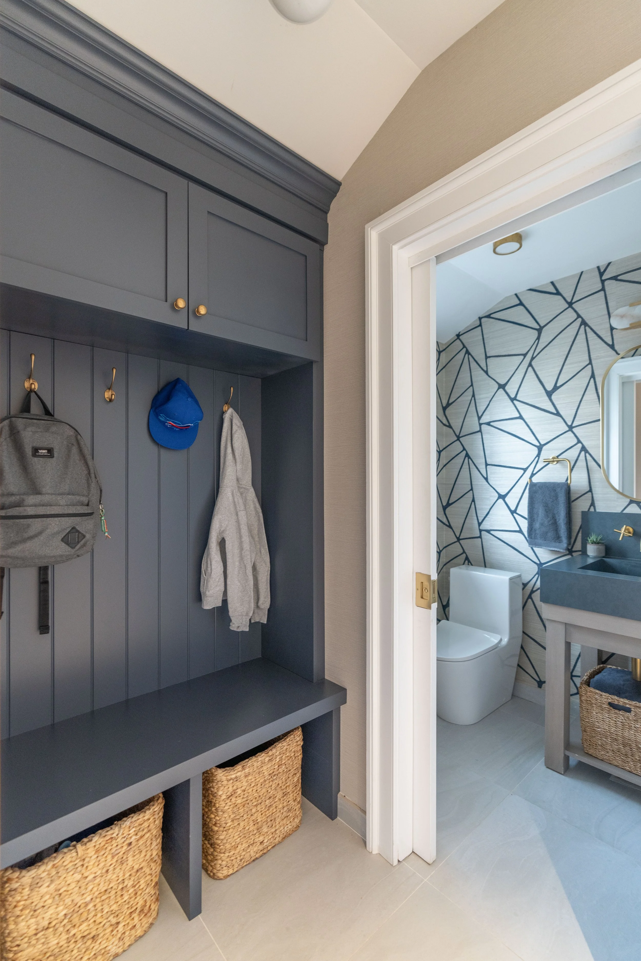 View of a home entrance with a blue coat rack wall with hooks holding a backpack, a blue cap, and a gray hoodie. There are two woven baskets underneath the coat rack, and part of a bathroom is visible with a toilet, a gray sink, a mirror, and a patte