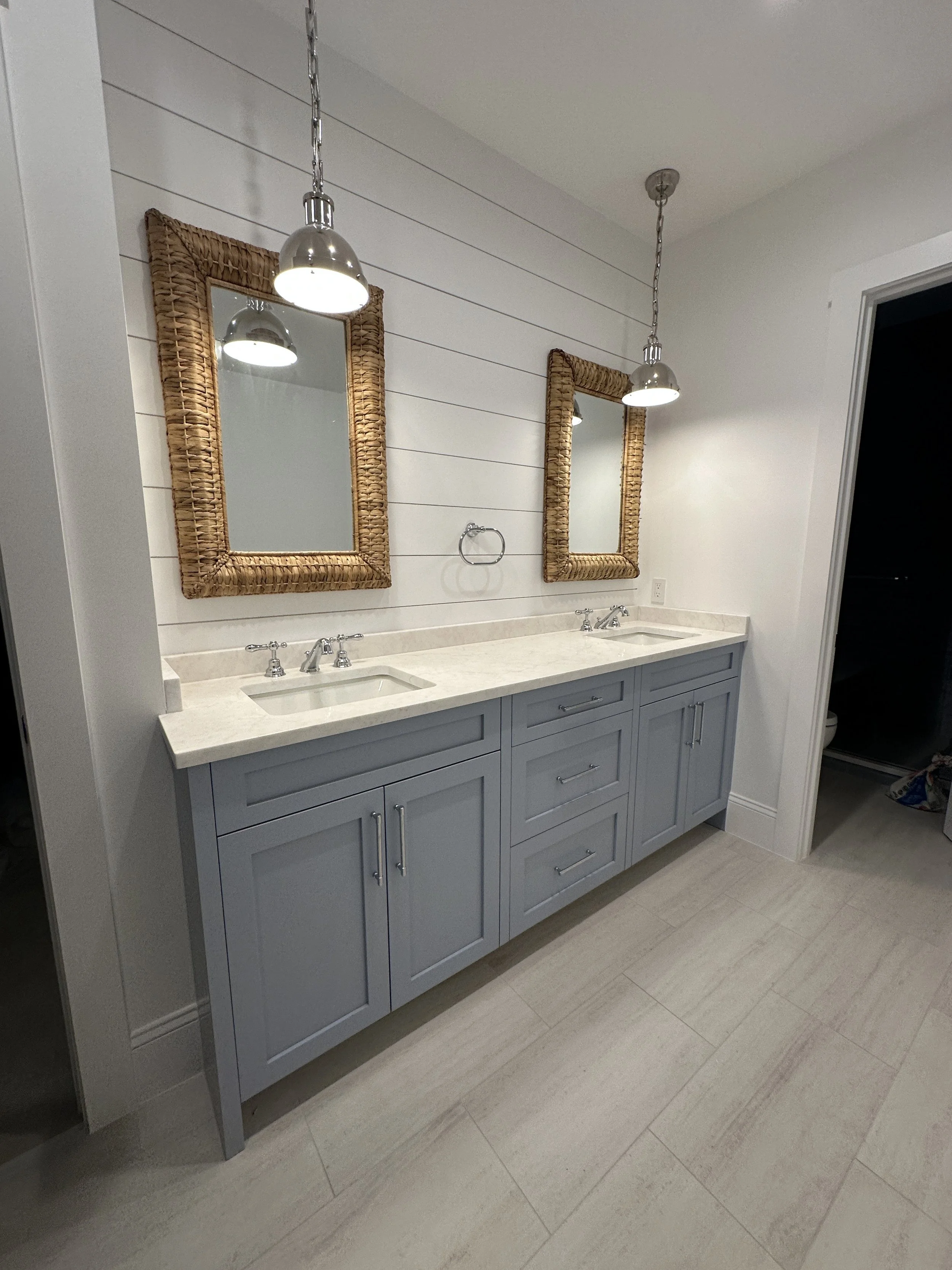 Double sink bathroom vanity with gray cabinets, white countertop, two rectangular mirrors with woven rattan frames, two pendant lights hanging above, white shiplap wall, towel ring, and a doorway to a dark-colored room.