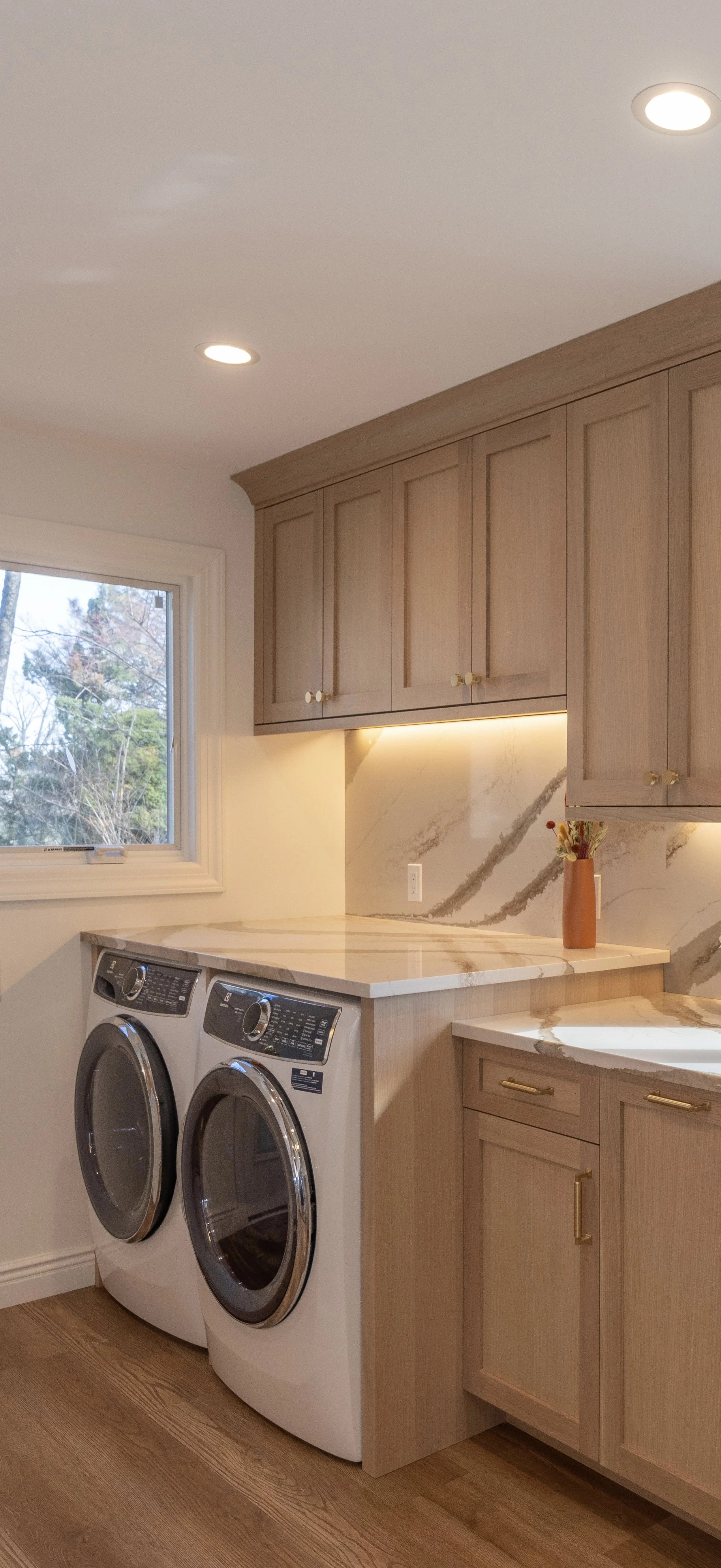 A laundry room with a window, wooden cabinets, a marble countertop, and a pair of front-loading washing machines.