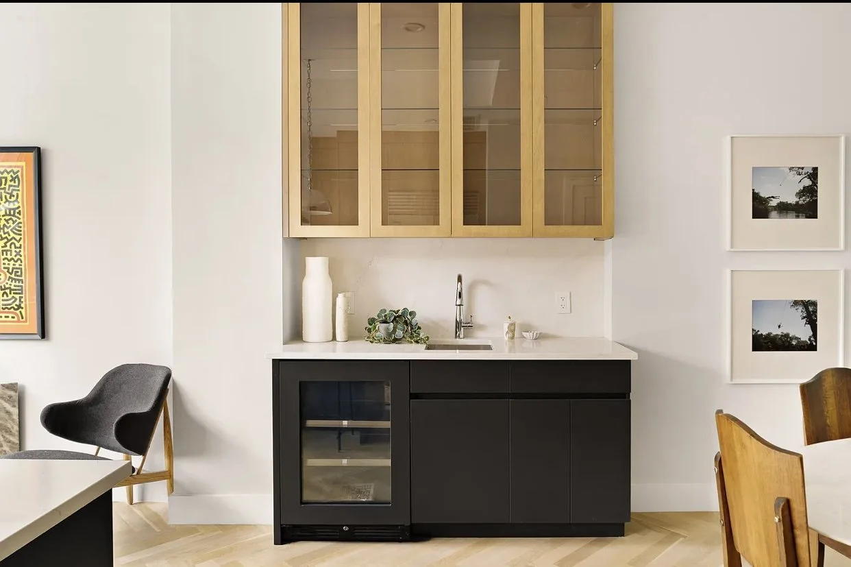 Modern mini bar area with black lower cabinets, a white countertop with sink, gold upper cabinets with glass doors, and decor including vases and plants. Part of a living or dining room with artwork and chairs.