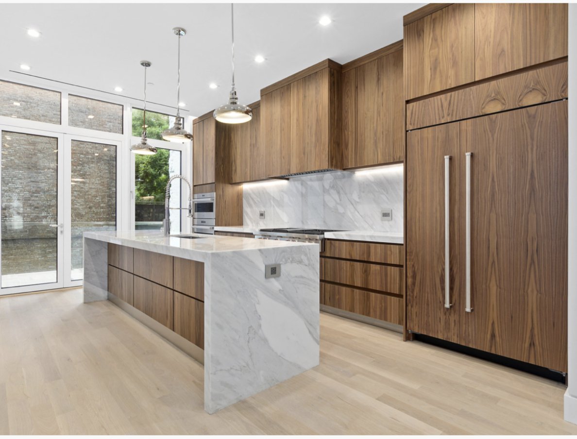 Modern kitchen with wooden cabinets, white marble island with gray veining, built-in oven, and large windows.