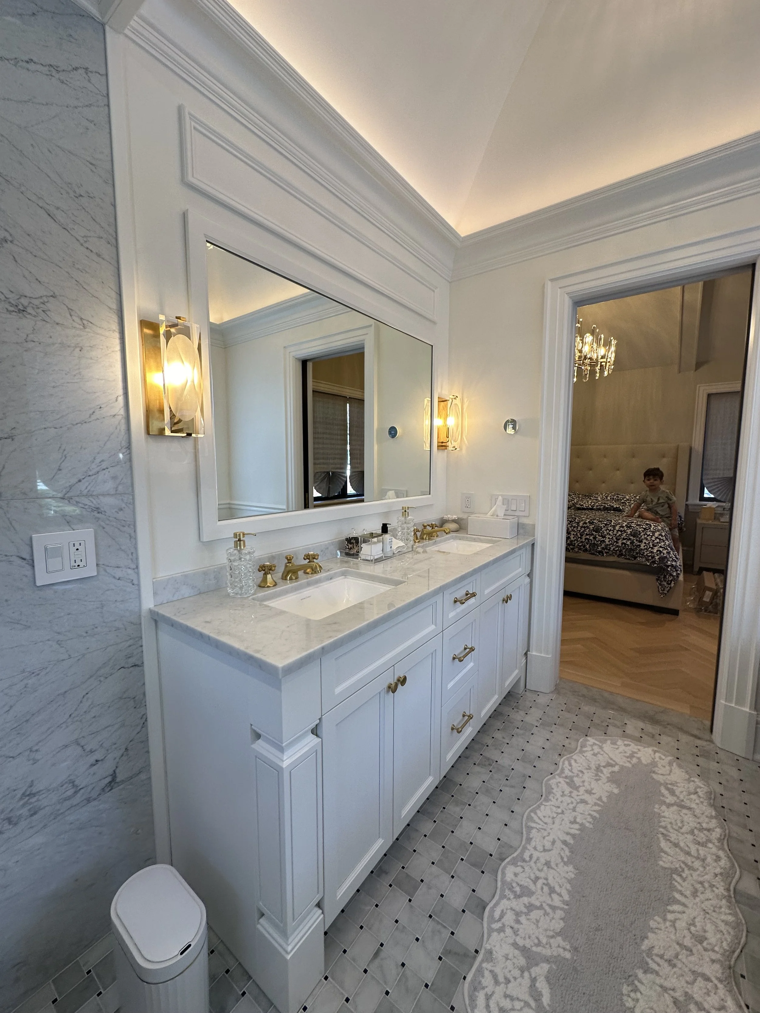 A luxurious bathroom with a double sink vanity, marble countertop, large mirror, gold fixtures, decorative soap dispensers, a small storage box, wall-mounted lights, and a patterned rug. Seen through an open doorway into a bedroom with a bed, chandelier, and a boy sitting on the bed.