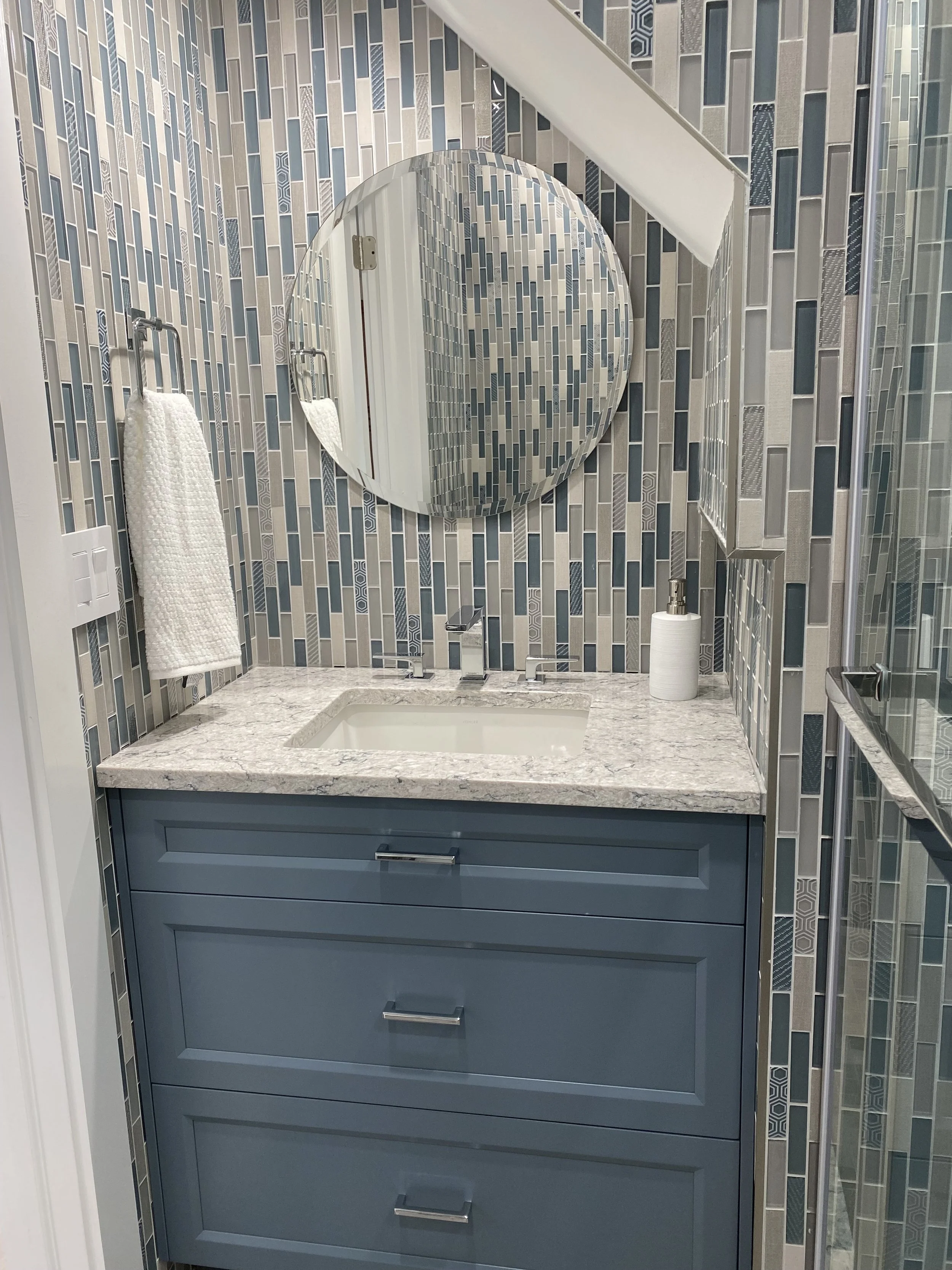 A bathroom with a blue vanity, marble countertop, and a round mirror above the sink. The wall features mosaic tile with shades of blue, gray, and white. There is a towel hanging on a hook to the left of the mirror and a soap dispenser on the right of the sink.