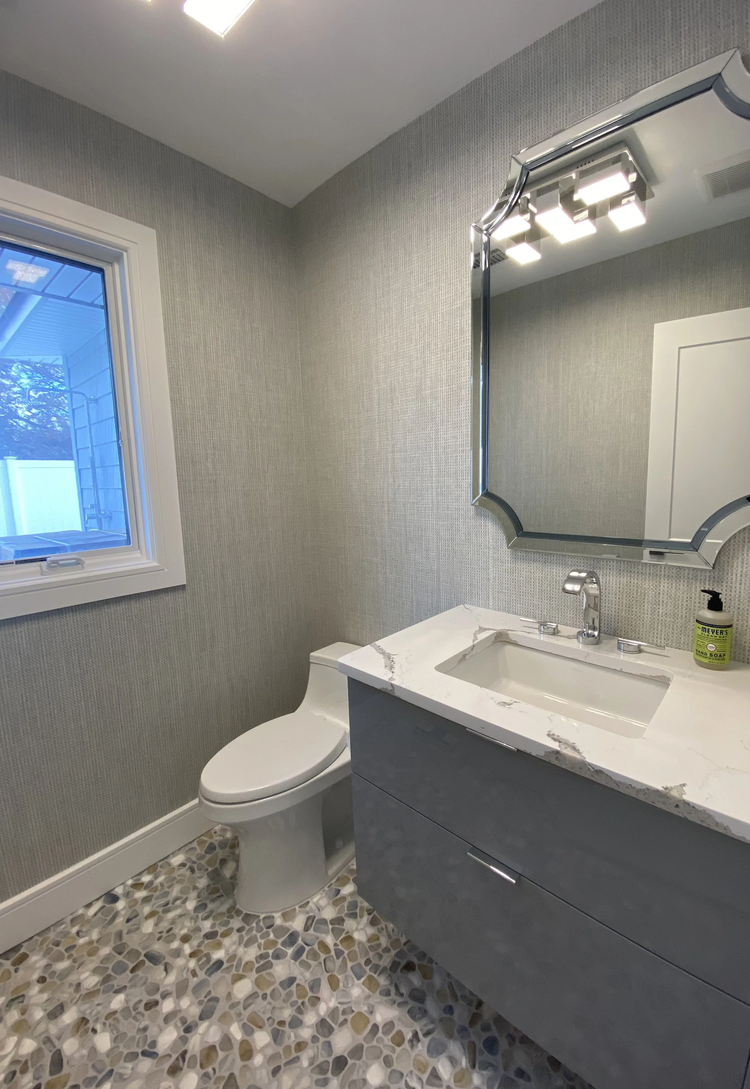 A modern bathroom with a small window, a white toilet, a gray vanity with a marble countertop, a rectangular sink, a silver faucet, a decorative mirror, and a green soap dispenser. The floor has pebble-style tiles.