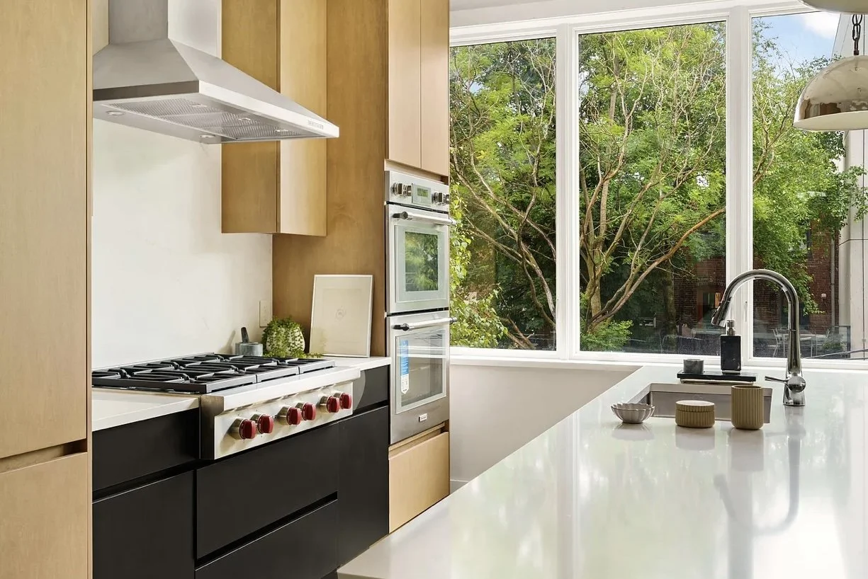 Modern kitchen with white countertops, black lower cabinets, and light wood upper cabinets. A window offers a view of green trees outside. There is a stove with red knobs, a double oven, a stainless steel vent hood, a faucet over a white sink, and de