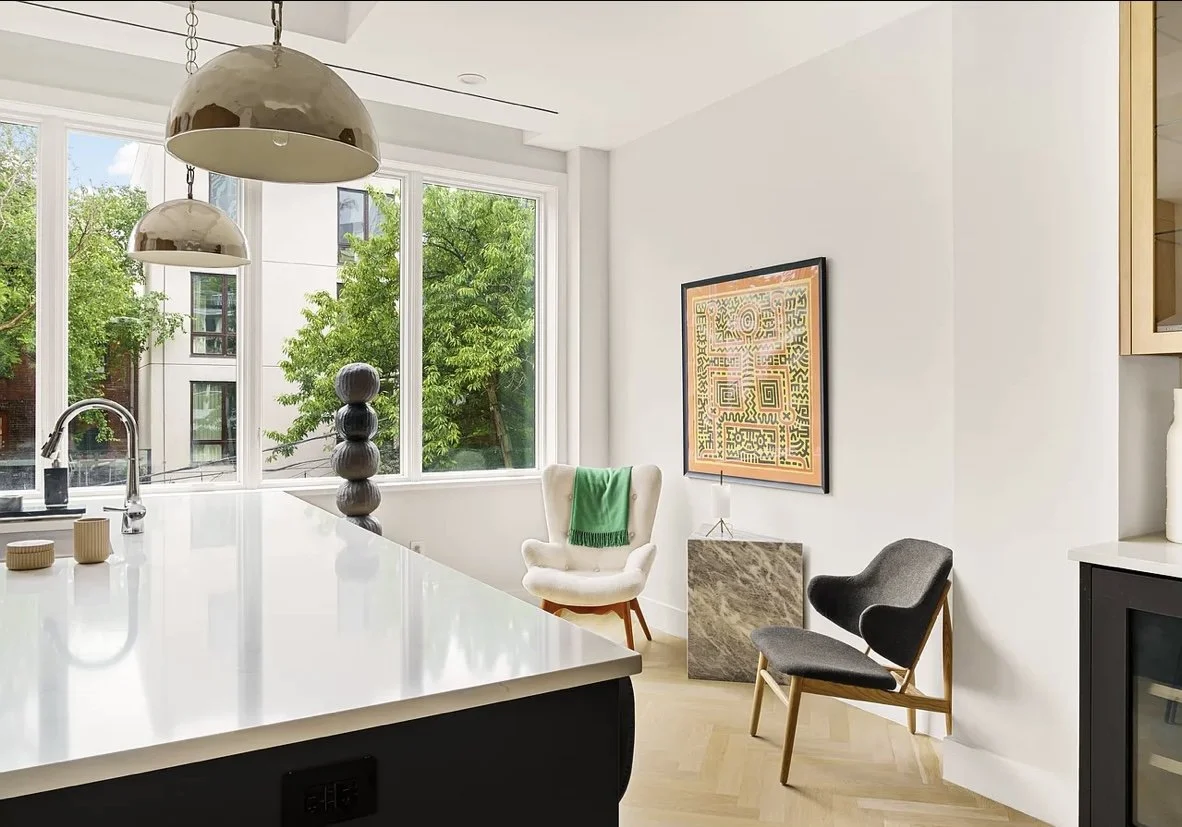 Modern kitchen and living area with large windows, a white counter, decorative objects, a white armchair with a green throw, a black and wooden chair, and colorful artwork on the white wall.