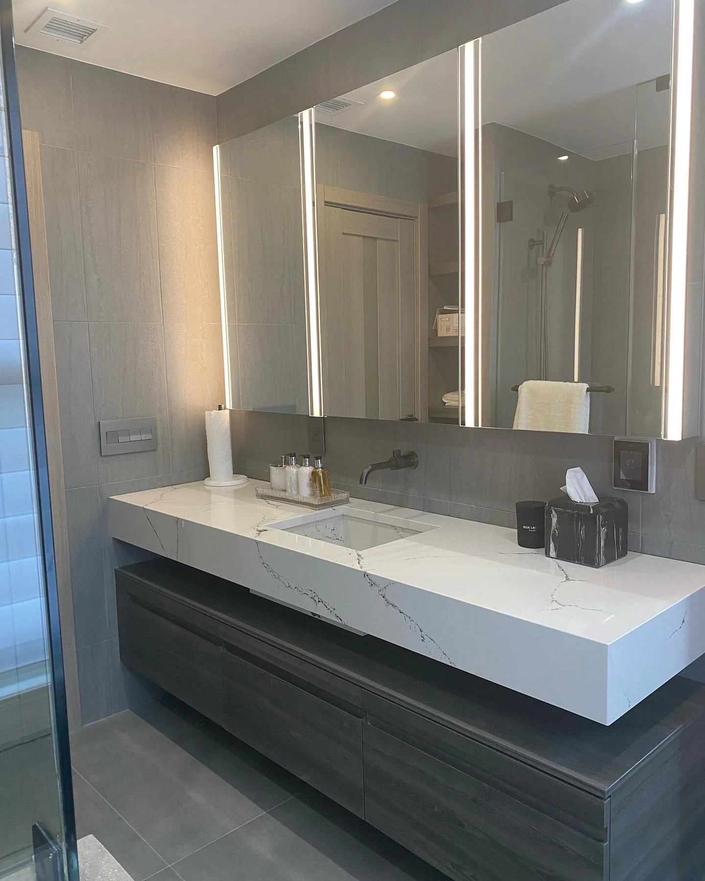 Modern bathroom with a large marble countertop, a built-in sink, and a large mirror with vertical lighting. There is a towel rack with a towel, soap bottles, and a tissue box.