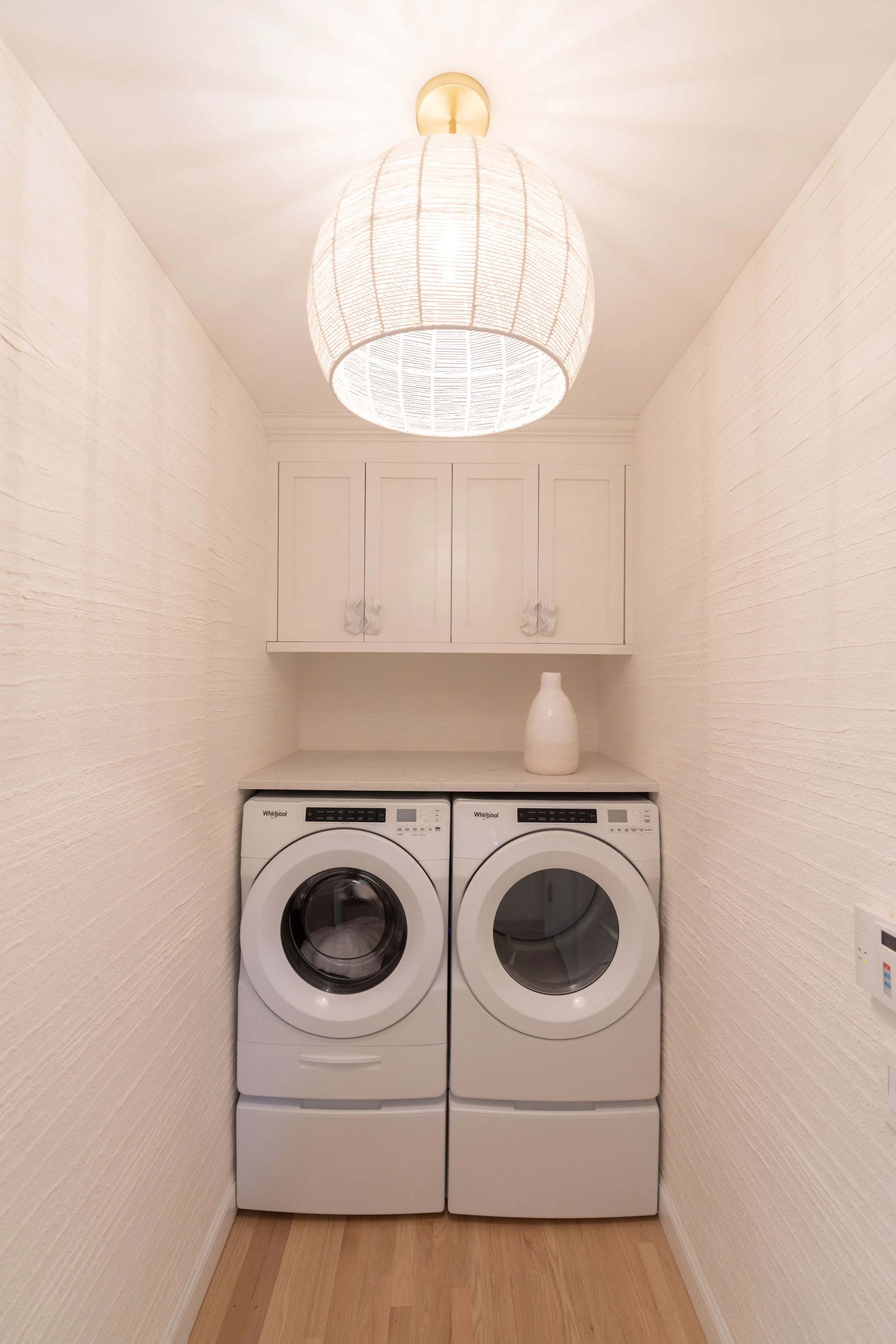 A laundry room with a washer and dryer, white cabinetry above, a white vase on the shelf, and a large ceiling light fixture.