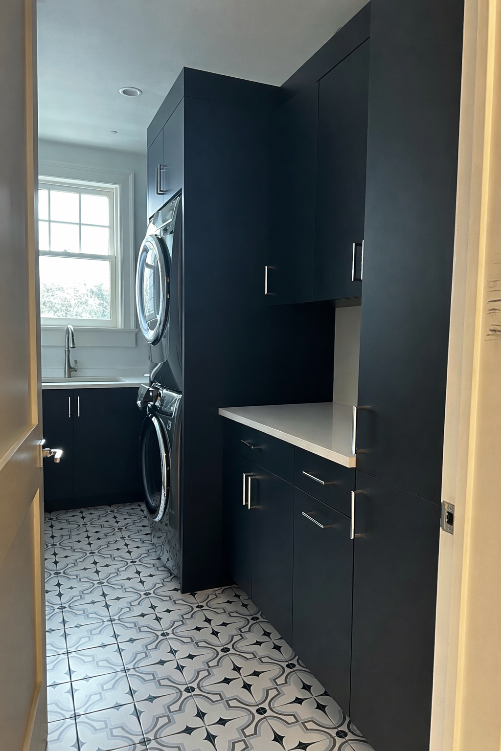 Laundry room with dark blue cabinetry, a white countertop, a window above the sink, and a stacked washer and dryer, with patterned floor tiles.