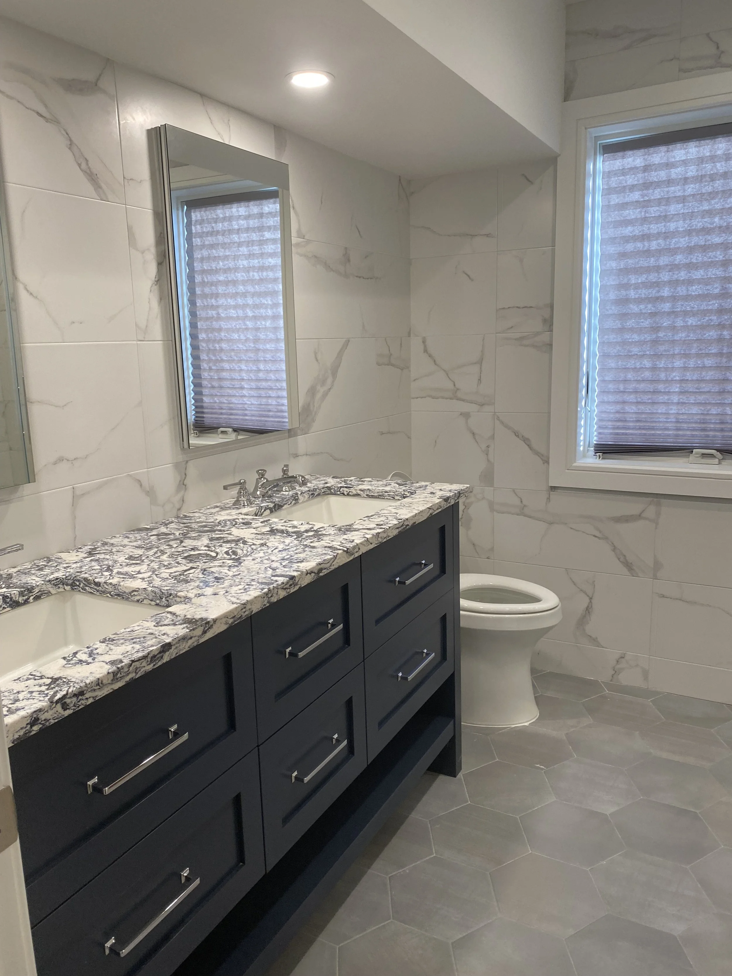 Modern bathroom with marble walls, gray hexagonal tile floor, navy blue vanity with granite countertop, twin sinks, and a mirror. There is a toilet and two windows with closed blinds.