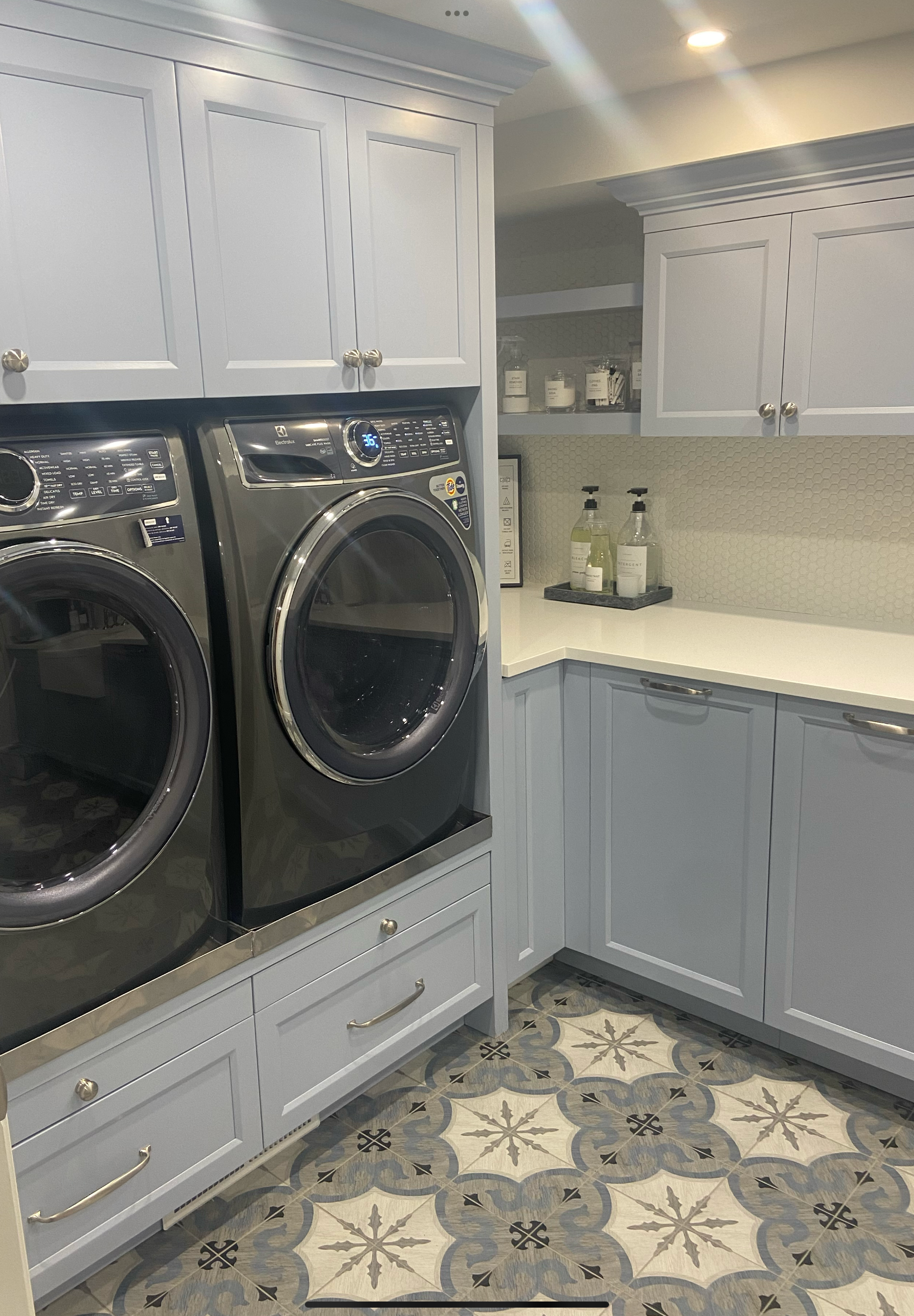 A laundry room with light blue cabinets, a front-loading washer and dryer, and a countertop with soap dispensers. The floor has patterned tiles in beige, gray, and black.