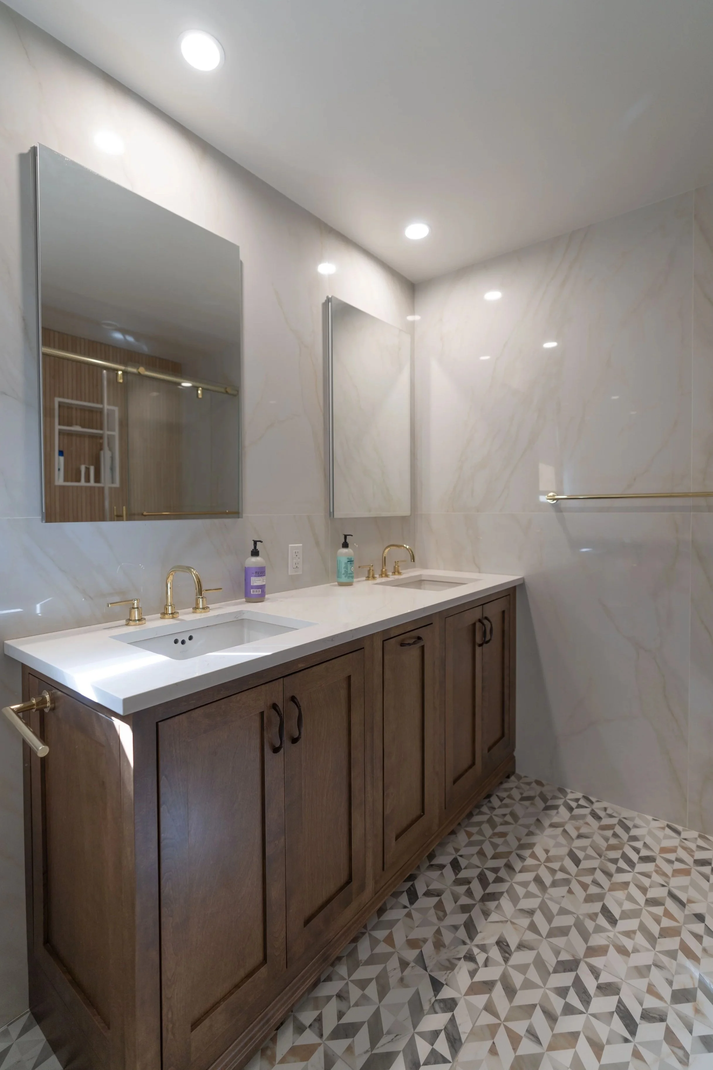 Modern bathroom with dual sinks, wooden vanity, large mirrors, marble walls, geometric patterned floor, and gold fixtures.