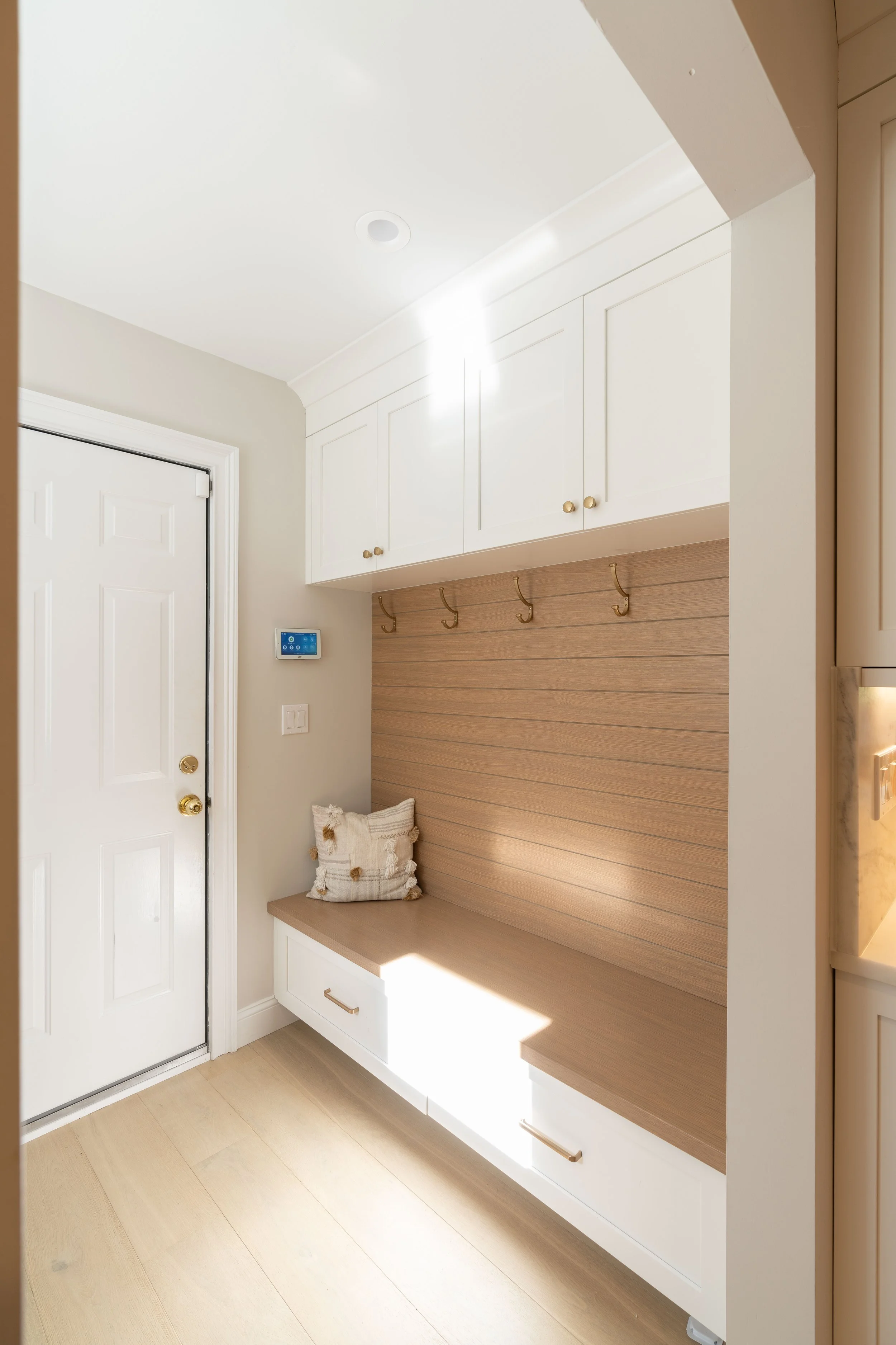 Entryway with white door, beige walls, built-in white cabinets and drawers, wooden bench with cushions, hooks for hanging items, and a small thermostat on the wall.