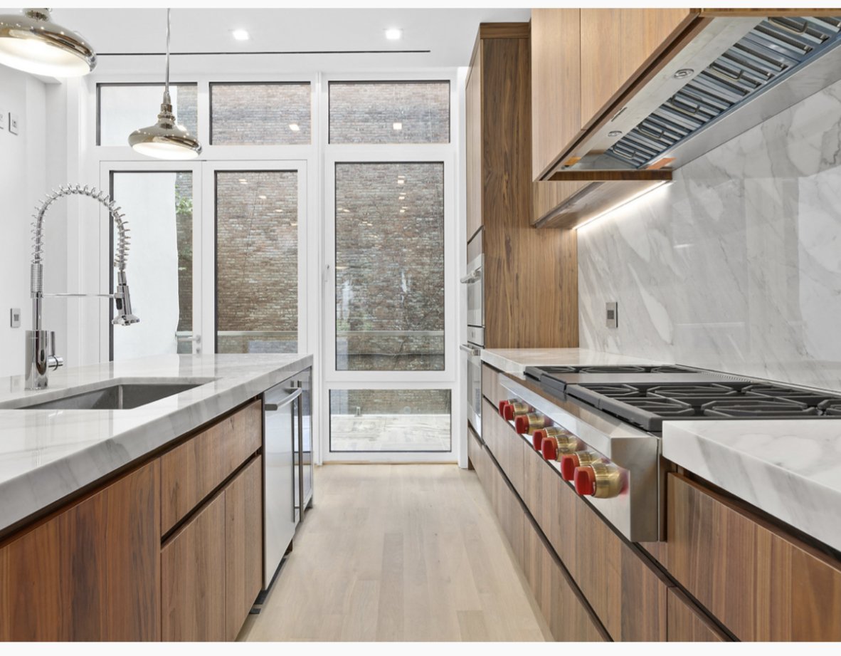 Modern kitchen with wooden cabinets, white marble countertops, stainless steel appliances, large windows, and a sliding glass door leading to an outdoor space.
