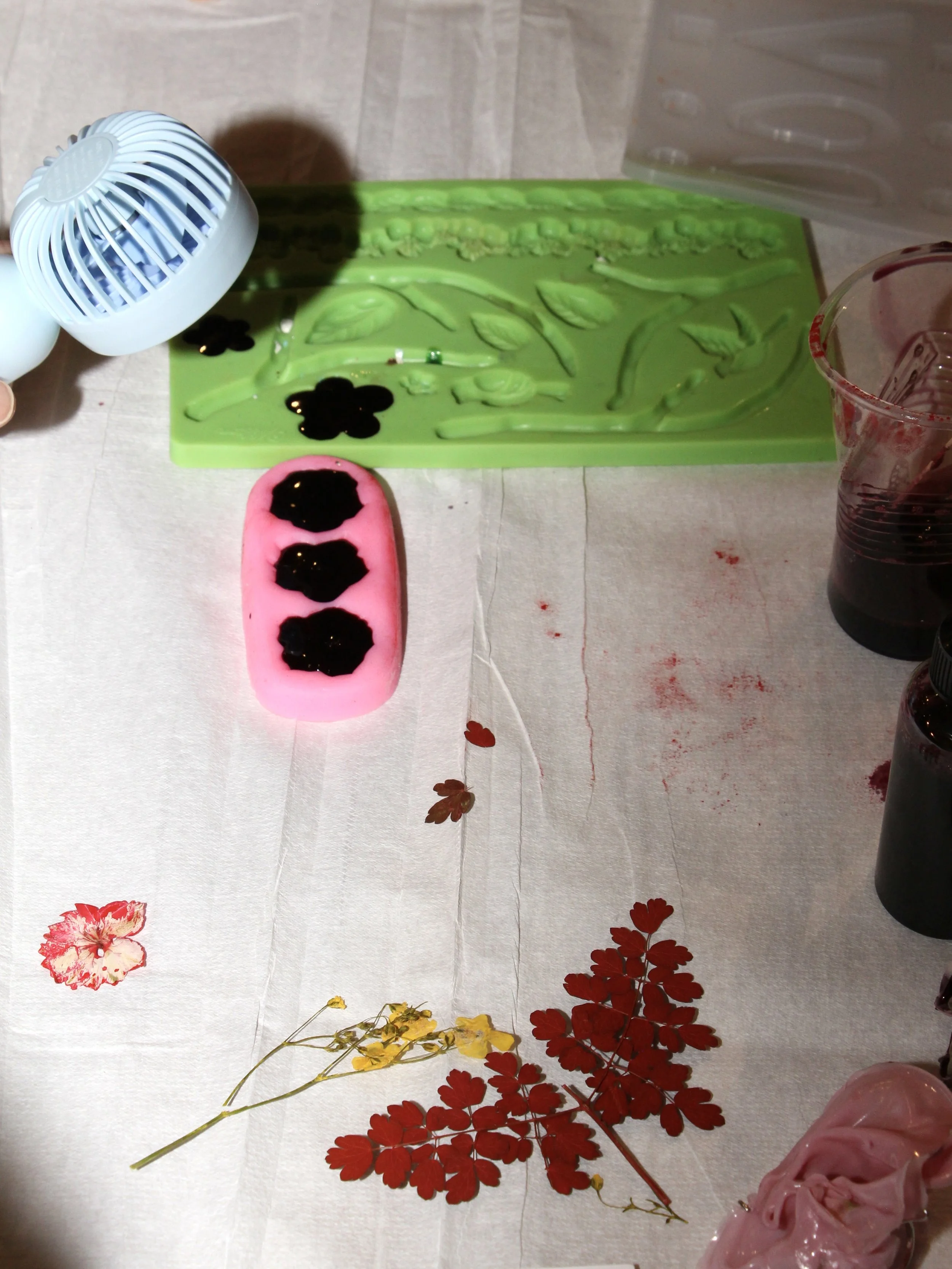 A table setup with a green silicone mold, a pink soap with black circle patterns, small jars of dark liquid soap or dye, a measuring cup, a small white fan, and decorative dried red and yellow flowers on a white tablecloth.