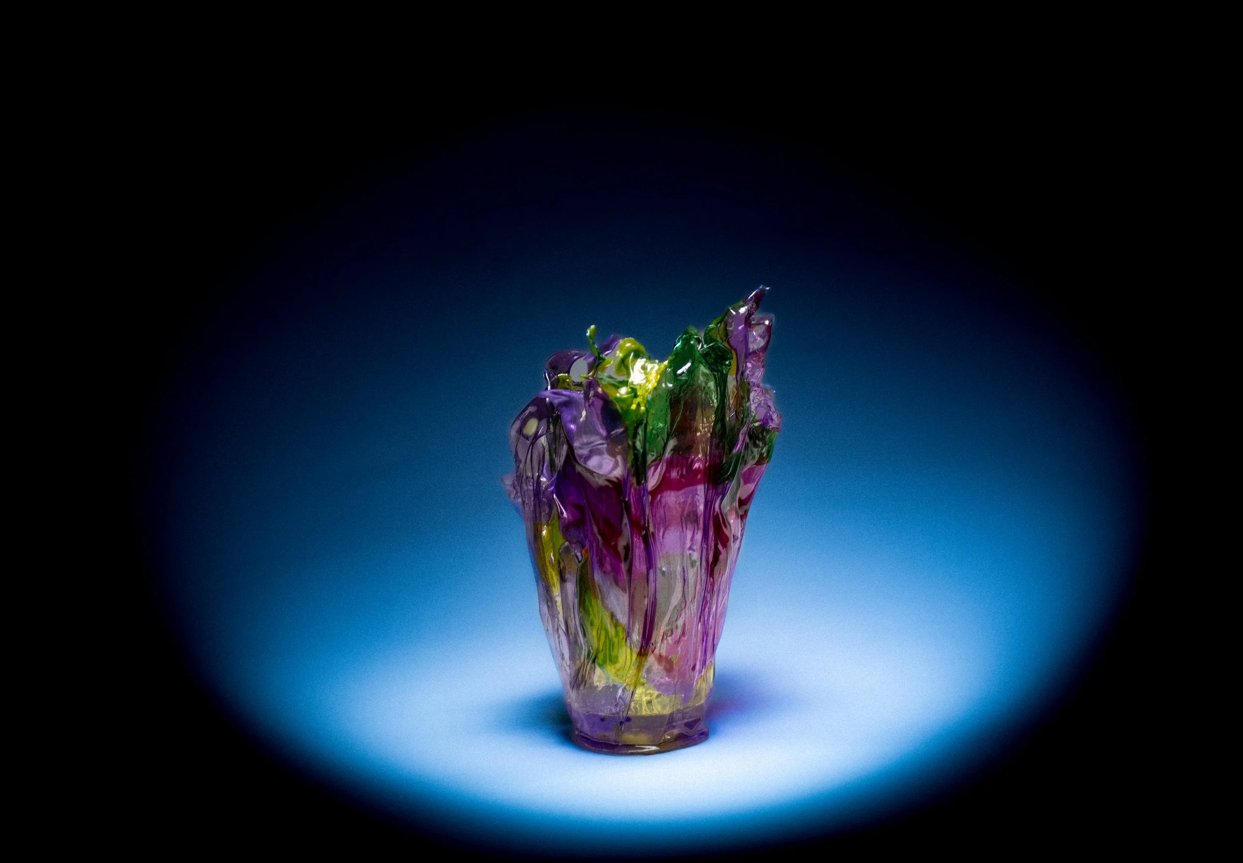 Colorful glass sculpture in a dark setting, illuminated by a spotlight, with purple, green, and pink hues.