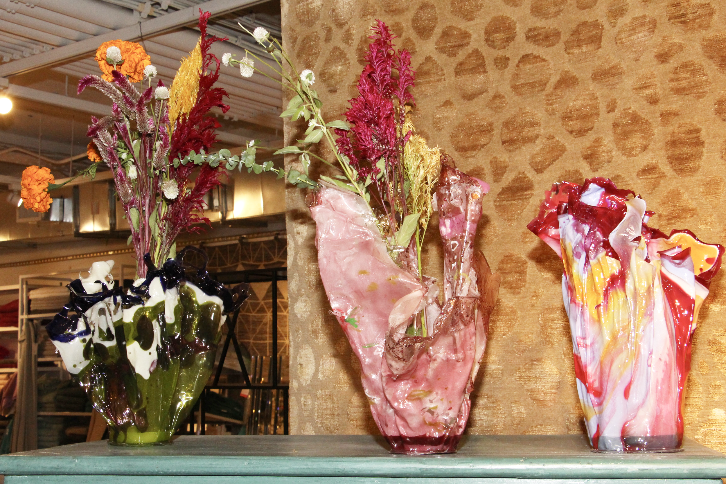 Three colorful glass vases with dried flowers on display against a textured wall.