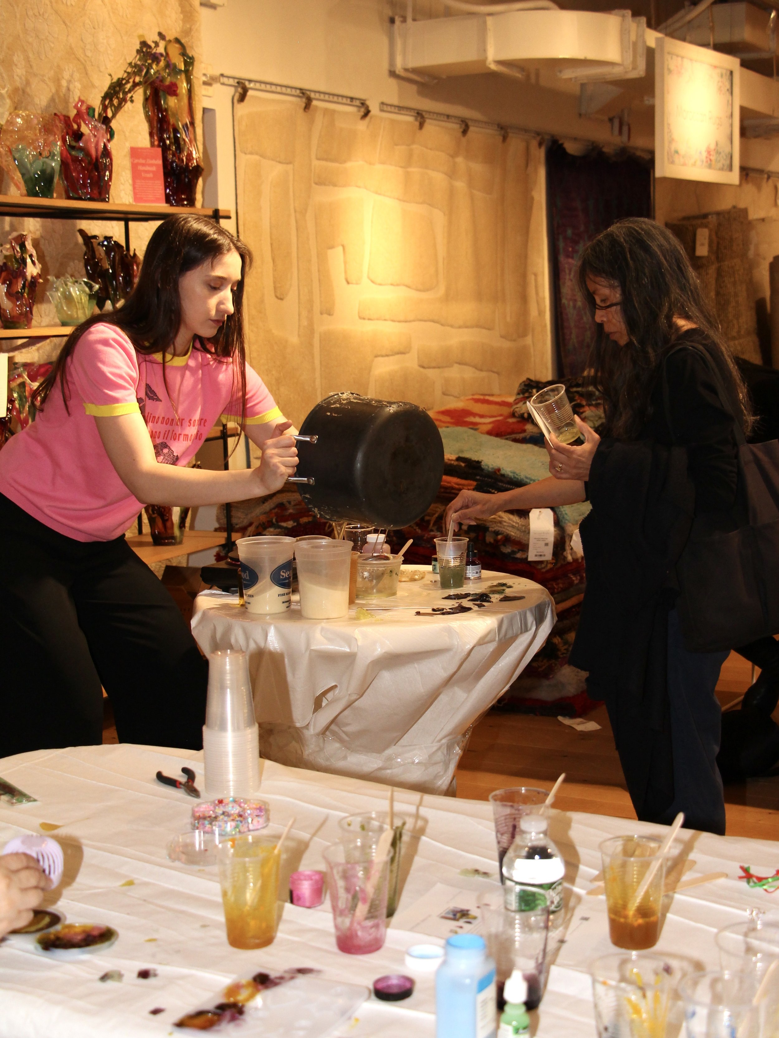 Two women engaged in a paint pouring activity at a craft table with various art supplies and drinks.