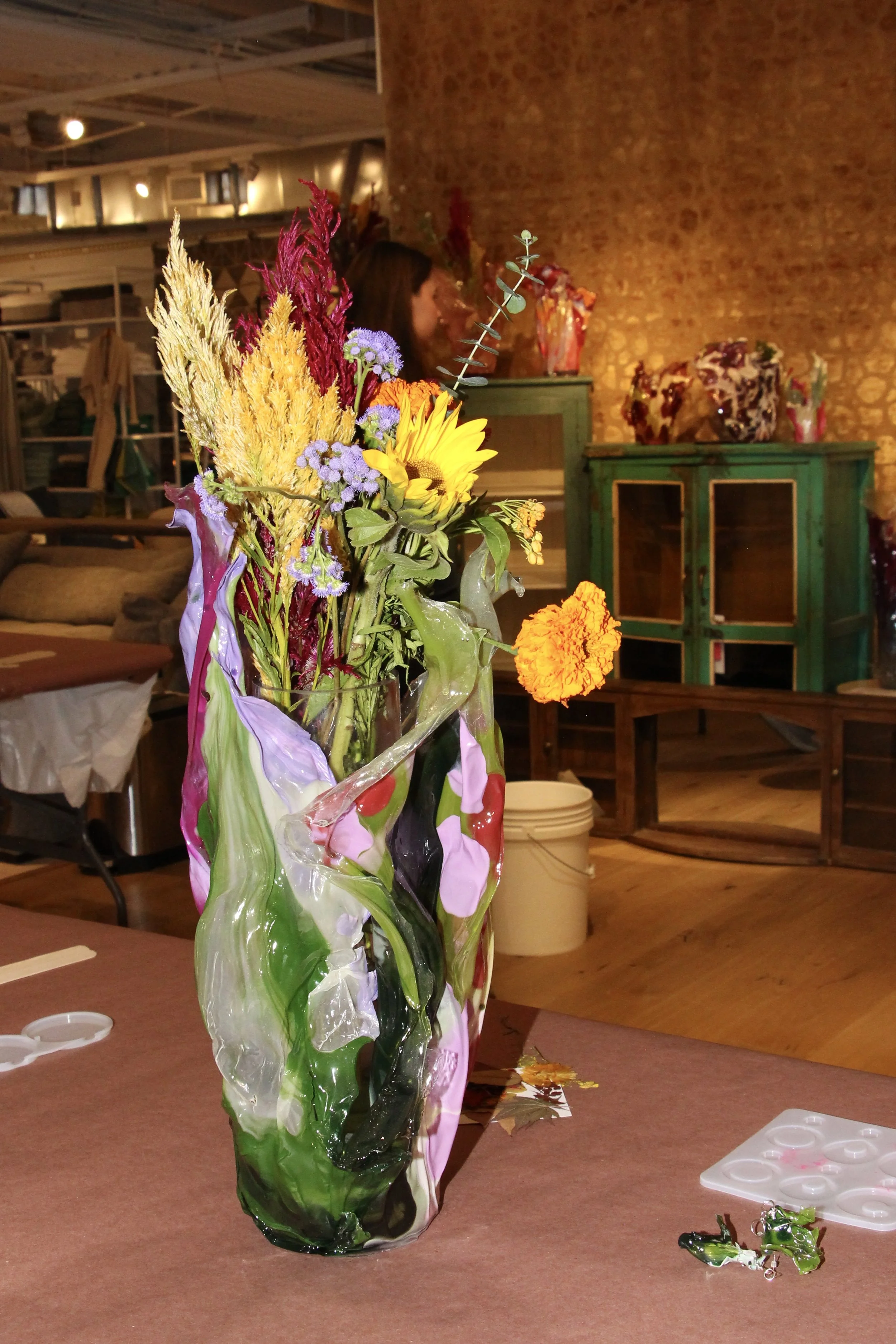 A colorful glass vase with a variety of dried flowers and greenery on a table in an indoor space with warm lighting, brick wall, and furniture in the background.