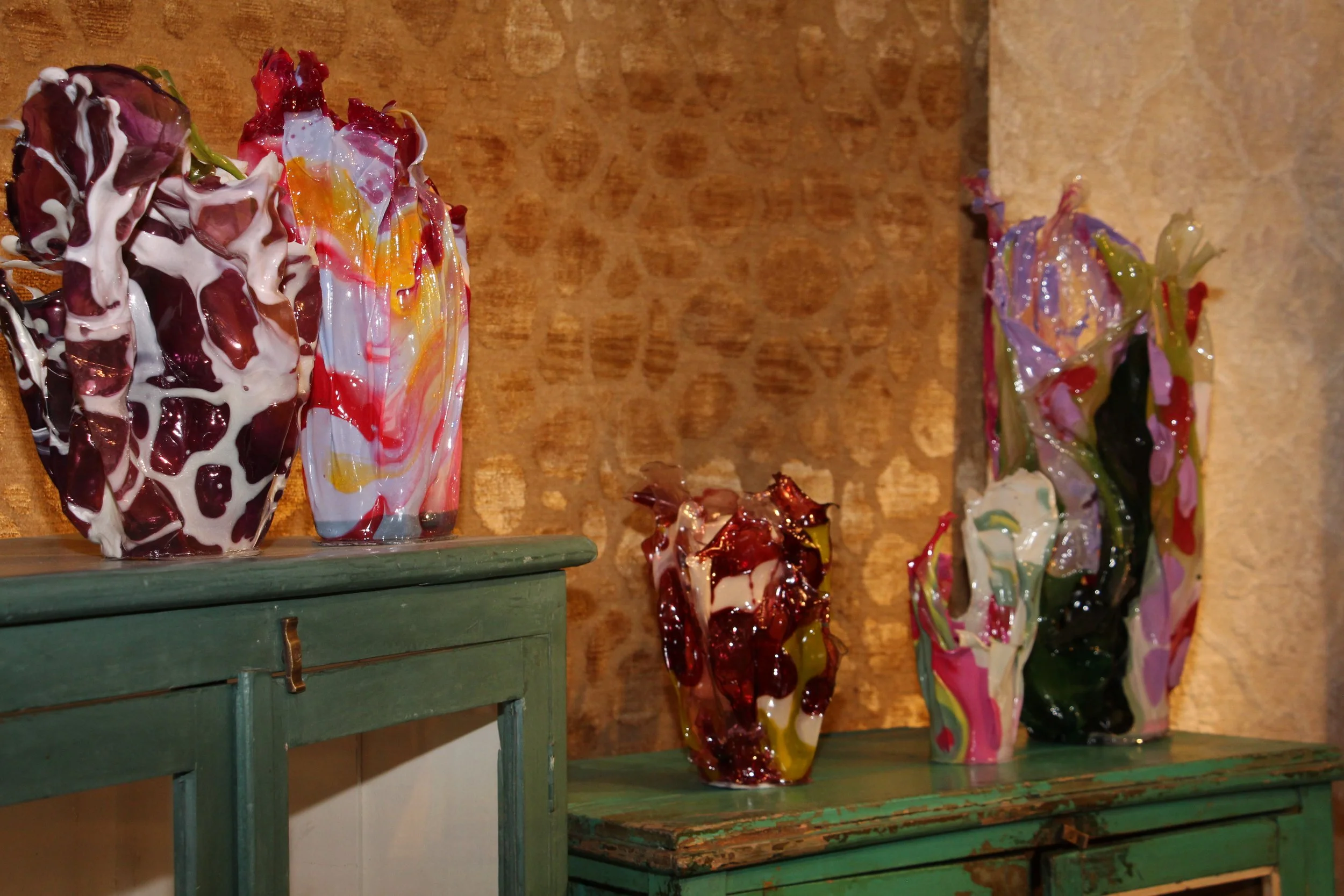 Four colorful abstract glass vases with swirling patterns and glossy surfaces displayed on rustic peacock green tables against a textured, ochre-colored wall.
