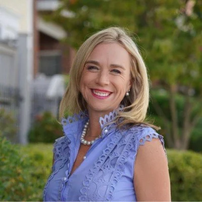A woman with blonde hair smiling outdoors, wearing a blue lace top and pearl necklace.