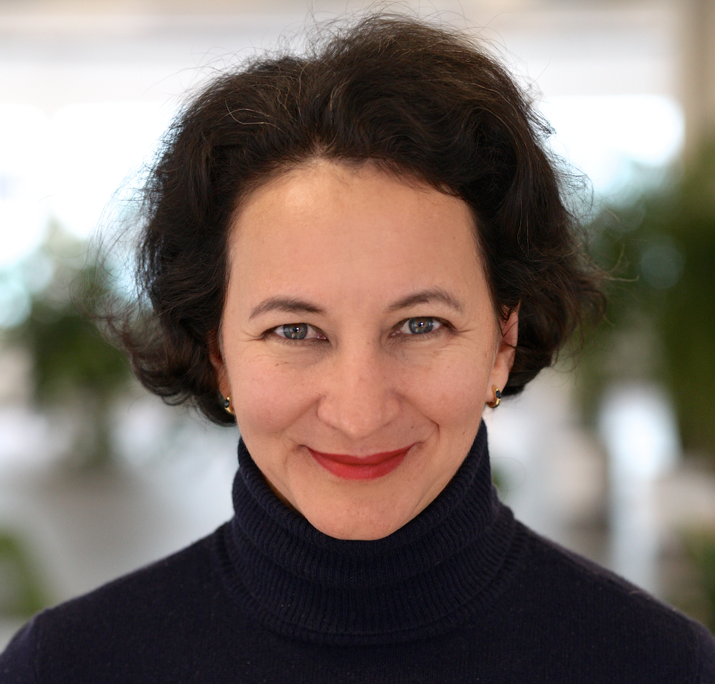 Close-up portrait of a smiling woman with dark curly hair, wearing a black turtleneck, with a blurred indoor background.