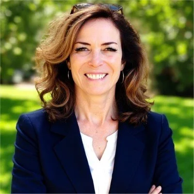 A woman with shoulder-length wavy hair smiling outdoors, wearing a dark blazer and white shirt, with a background of green trees and sunlight.