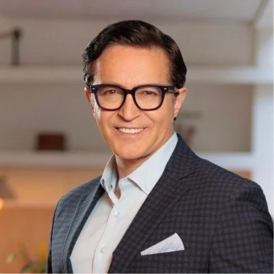 A man in a dark checkered suit jacket, white shirt, and an accident on his pocket, smiling, wearing black glasses, in an indoor setting.