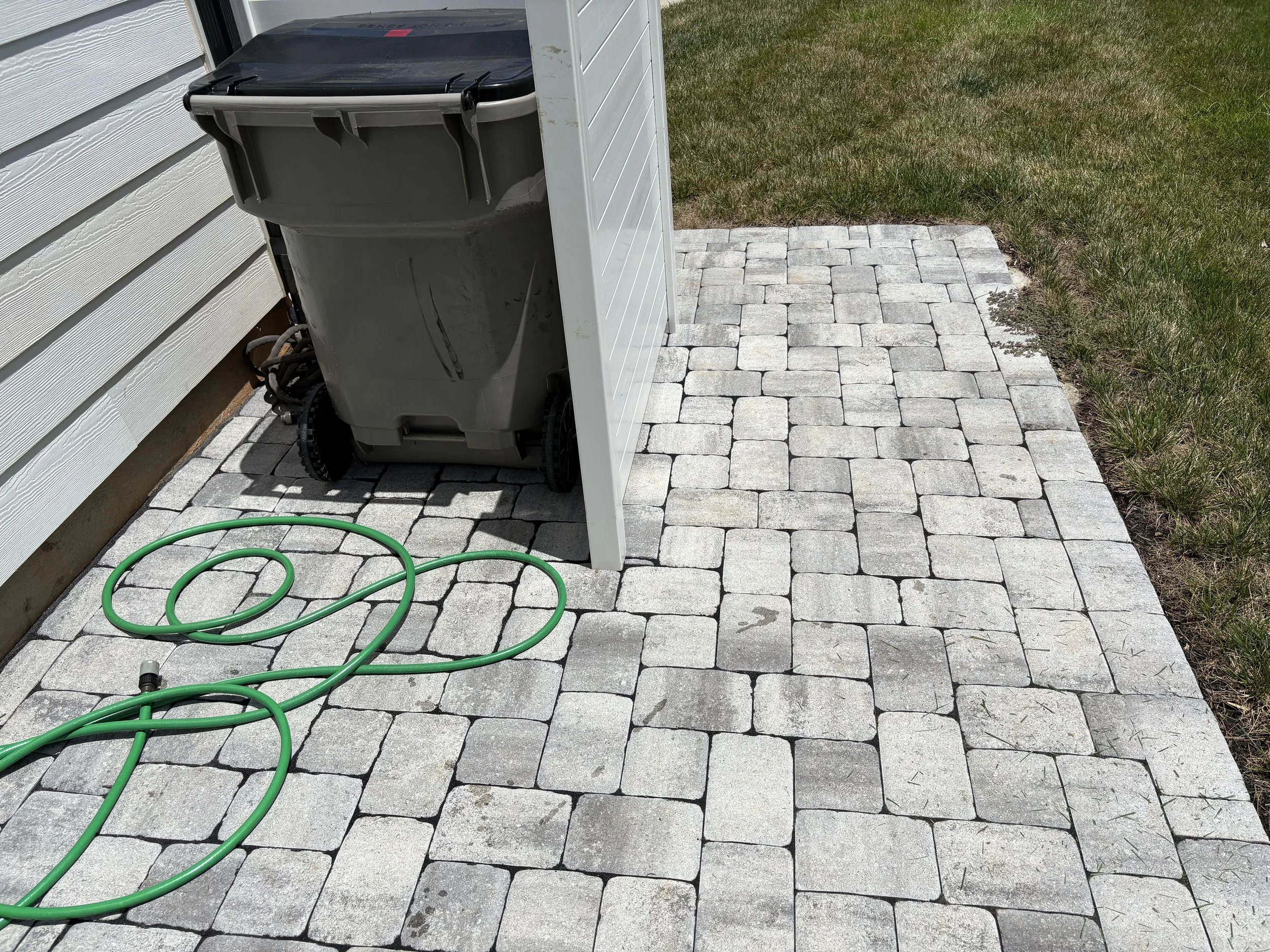 Backyard patio with grey stone pavers, a green garden hose, a trash bin, and a white vertical panel beside the bin, with grass on one side.