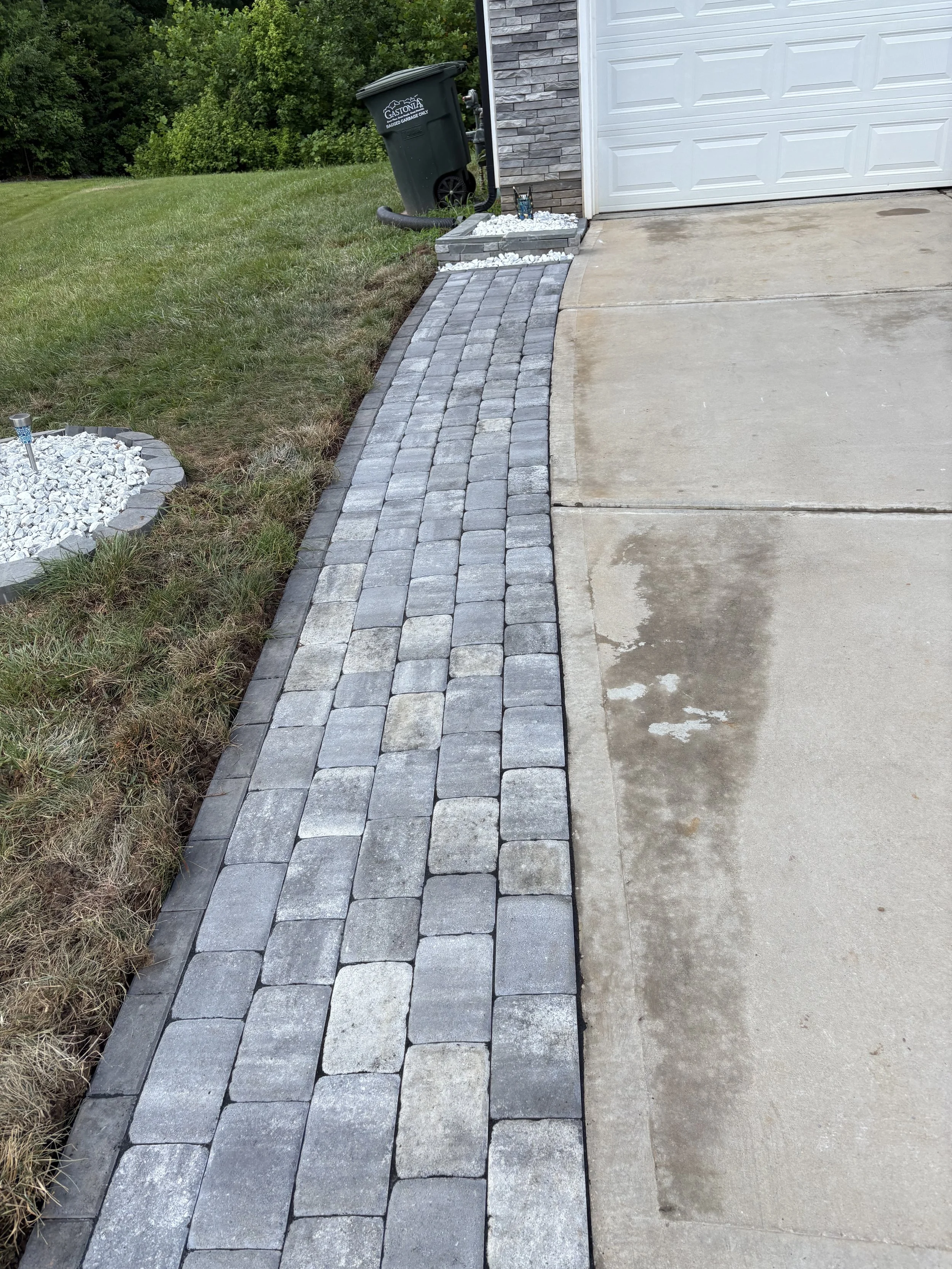Landscape with a curved gray brick walkway next to a driveway, with a grassy lawn and a stone border to the left, and a garage with a white door and gray brick exterior to the right.