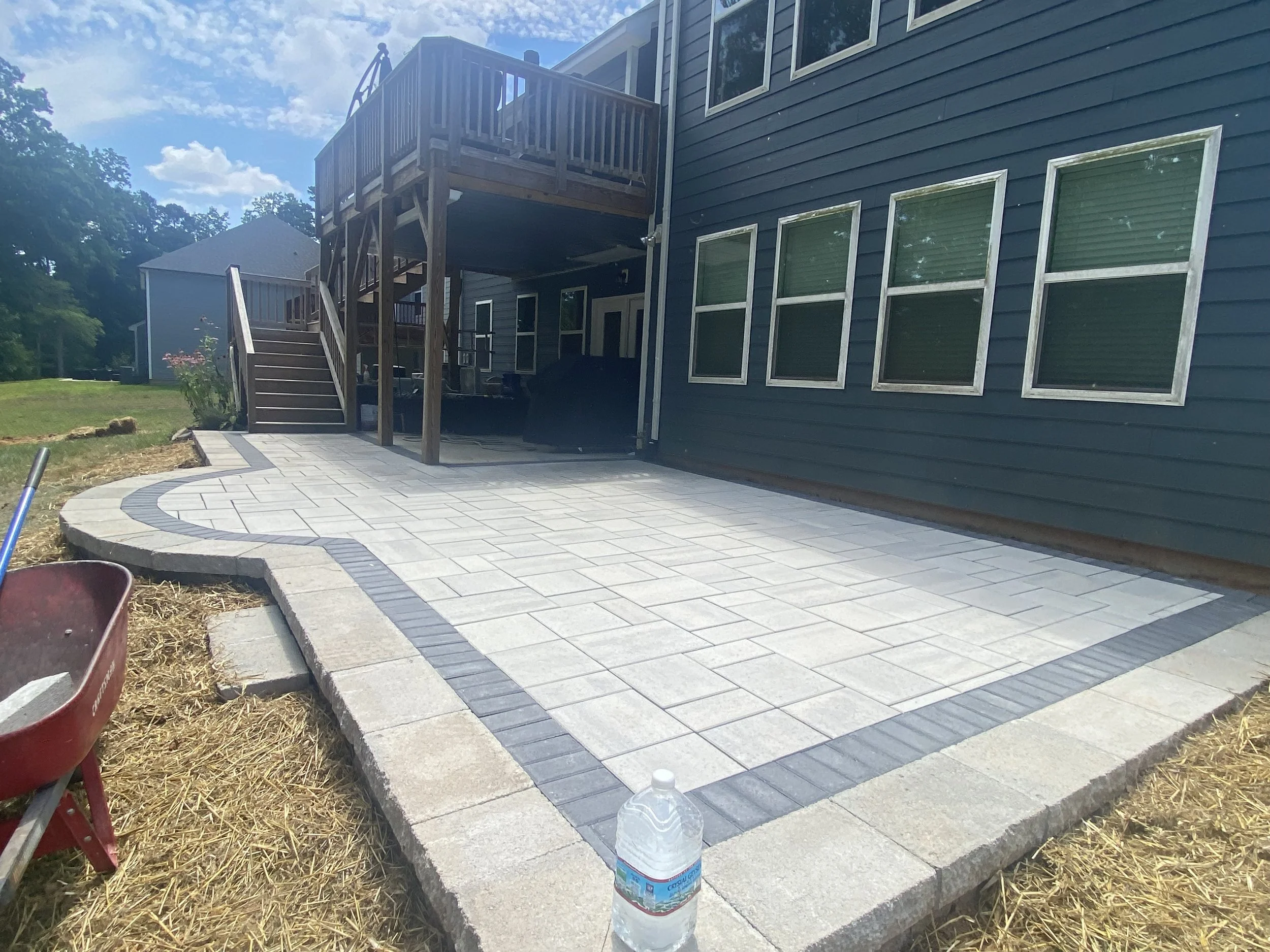 Newly paved backyard patio with light gray and dark gray tiles, a water bottle placed on the edge, and a red wheelbarrow on the grass.