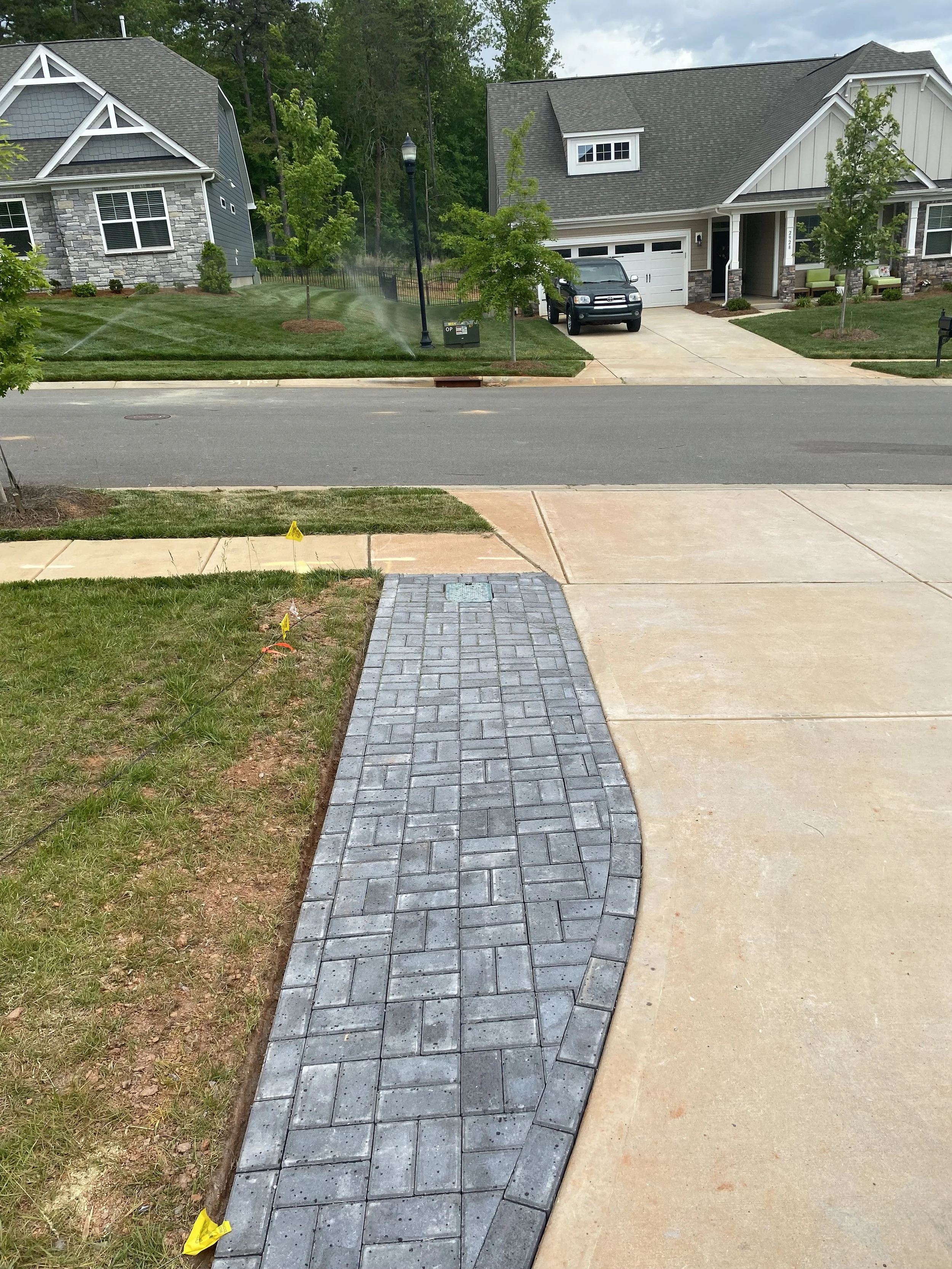 View of a residential neighborhood with a newly paved sidewalk, lawn, trees, and houses in the background. Spray irrigation is visible on the lawn of the house across the street.