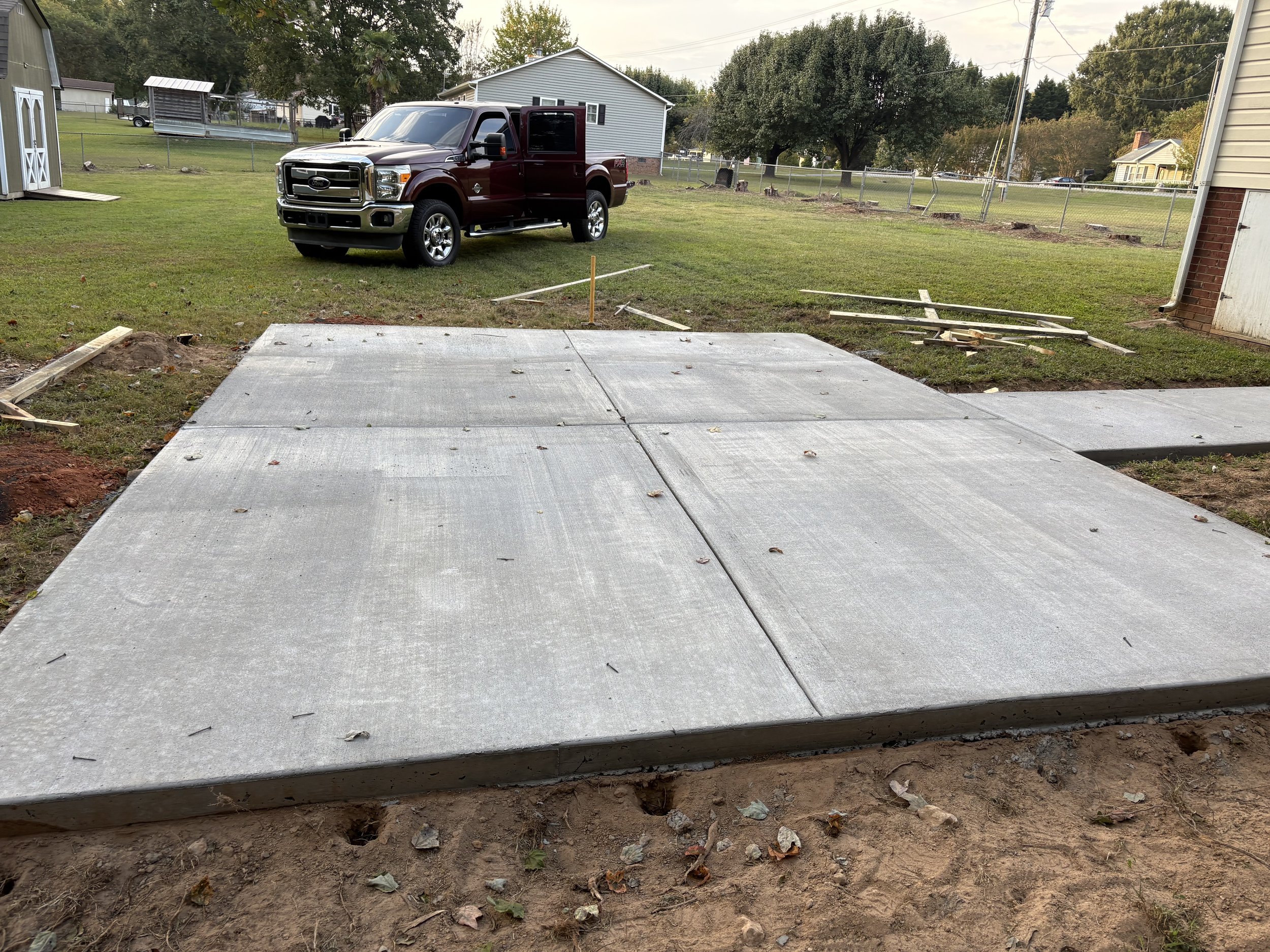 Concrete sidewalk being installed in a backyard, with a red truck parked in the grass nearby, and construction materials scattered around.