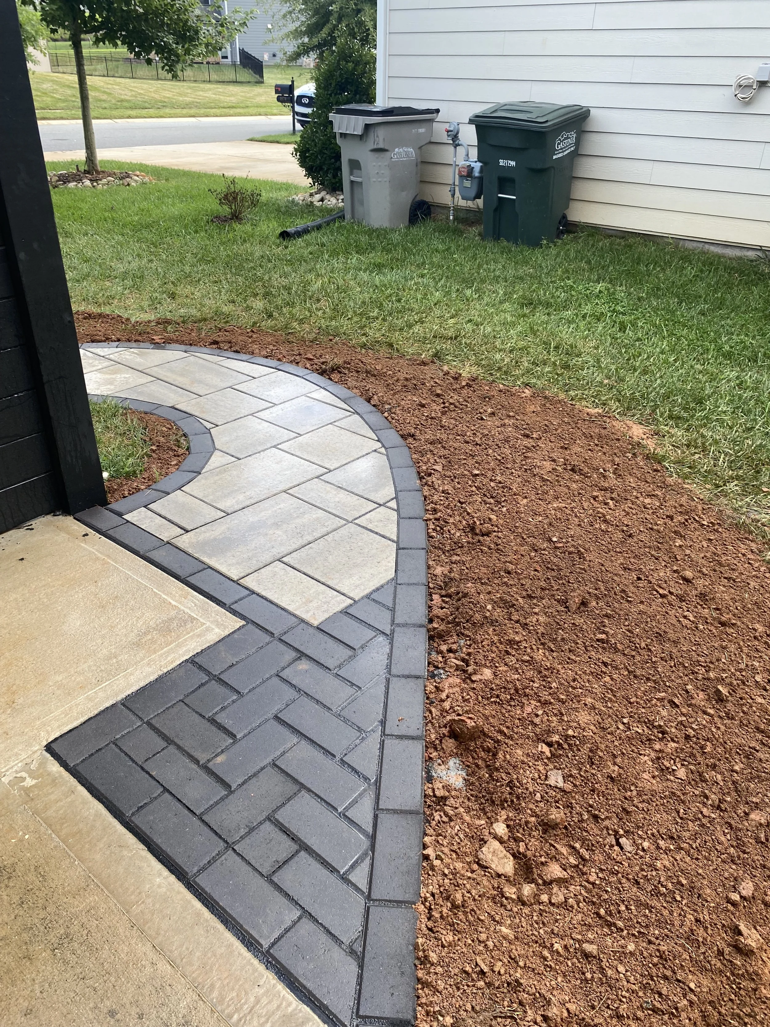 A newly constructed brick pathway with a curved design near a house, bordered by grass and soil, with two trash bins and a garden hose reel against the house wall in the background.