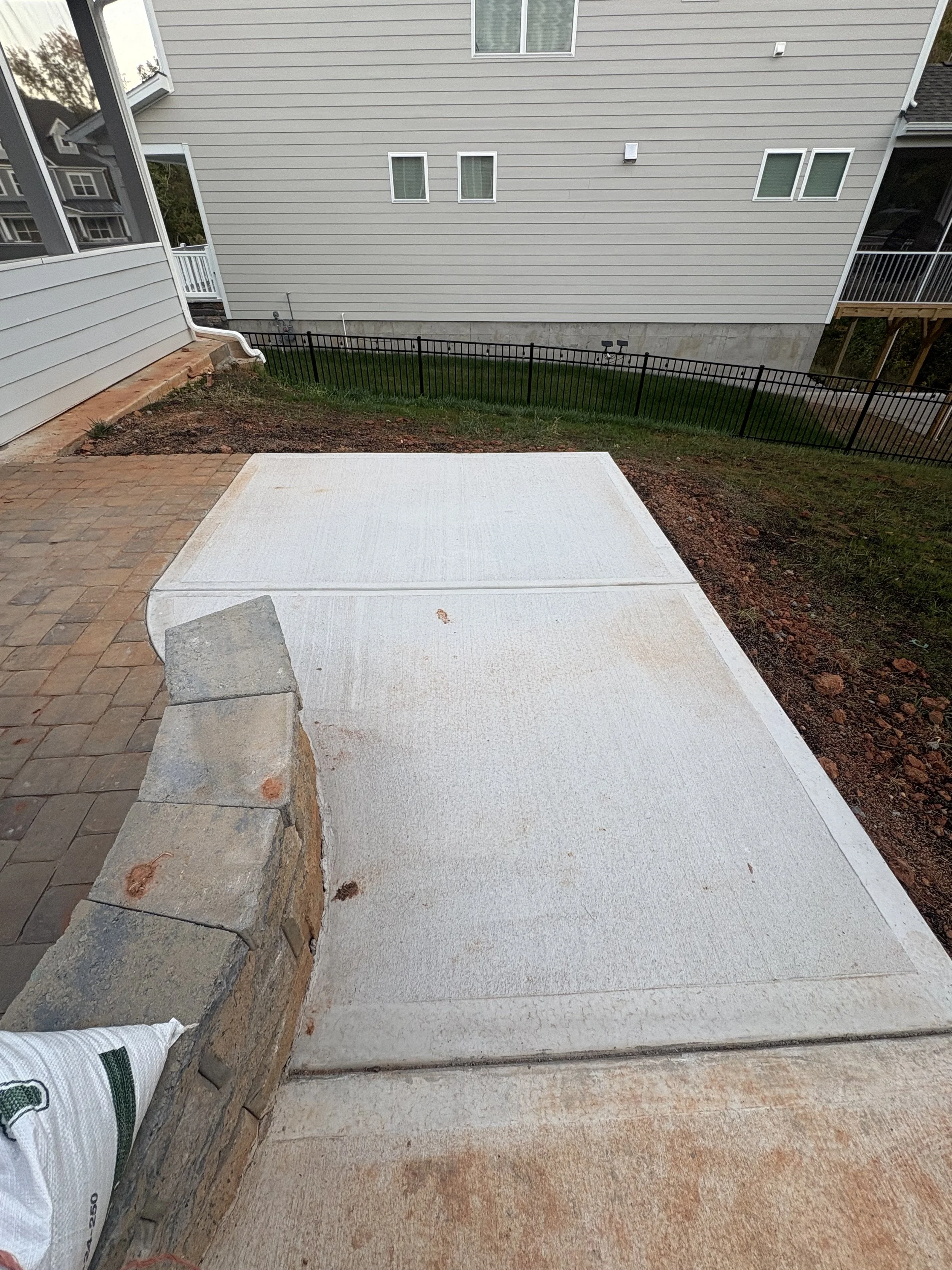 Newly poured concrete patio with a partial stone wall at the edge, next to a brick-paved section in a backyard with a house and fenced yard in the background.