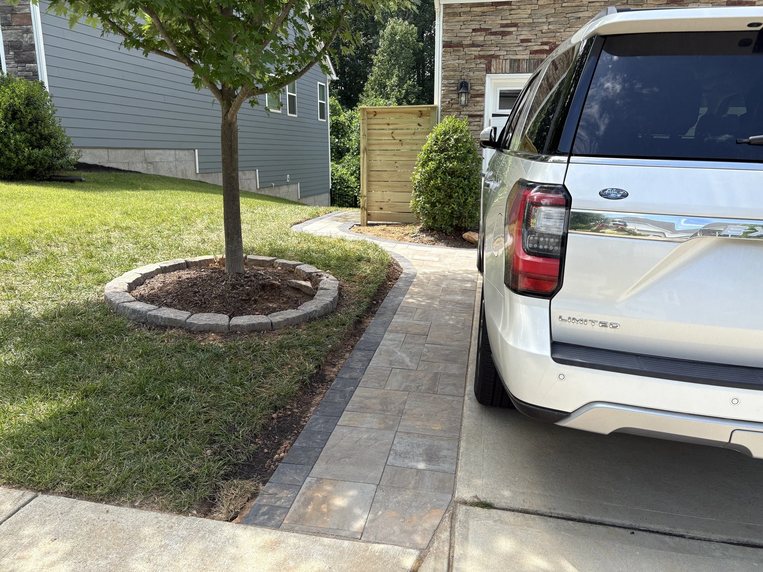 A suburban driveway with a white SUV parked next to a manicured lawn with a young tree in a stone-bordered mulch bed. The driveway leads to a house with brick and siding exterior, and a wooden privacy fence.
