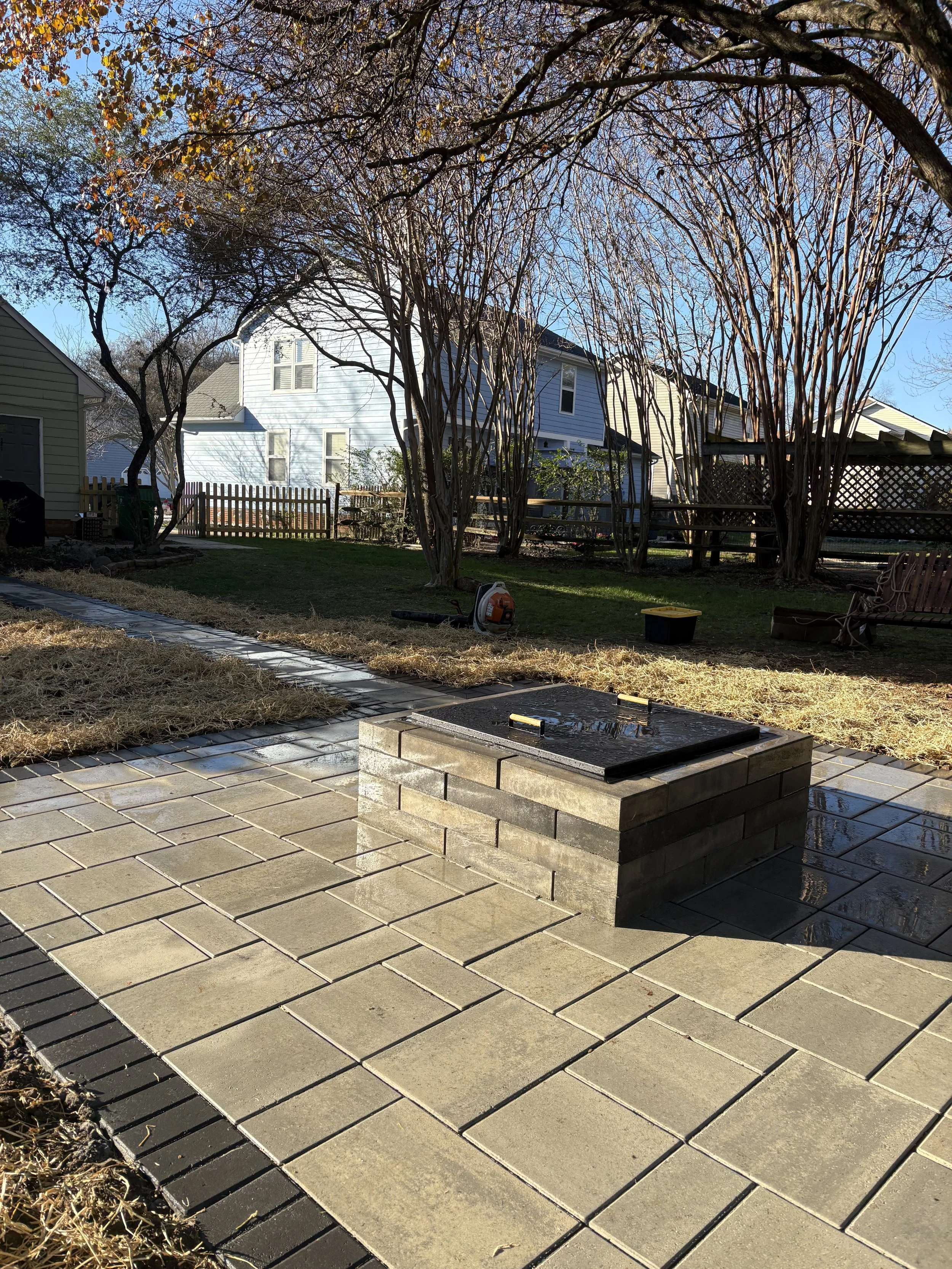 Backyard with newly paved stone patio, small brick fire pit with a black cover, leaf blower, bench, and a black container, surrounded by leafless trees and fenced yards.