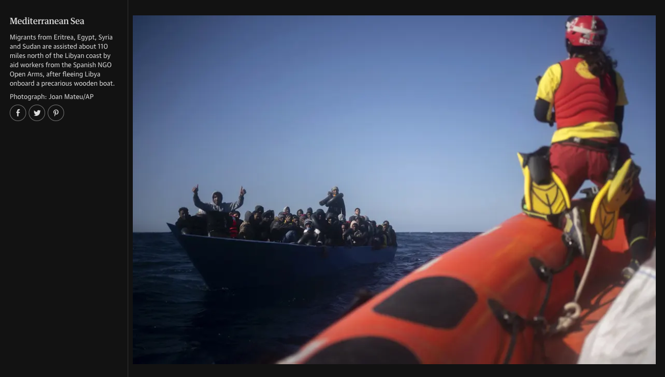 Rescue boat in the Mediterranean Sea with migrants from Eritrea, Egypt, Syria, and Sudan, being assisted after fleeing Libya on a wooden boat, with a rescue worker in the foreground on an orange inflatable boat wearing a helmet and red vest.