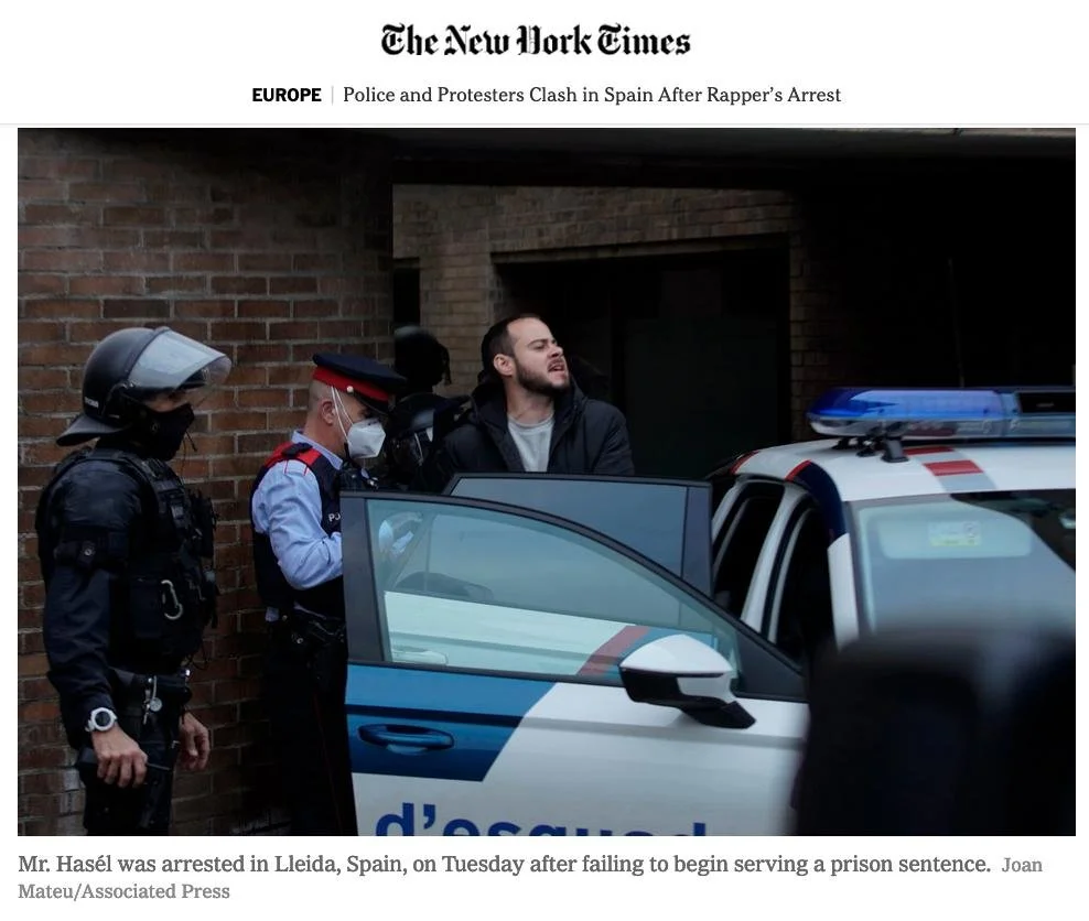 A man being detained by police officers, standing next to a police car with its door open. The scene is set against a brick wall in Lleida, Spain.