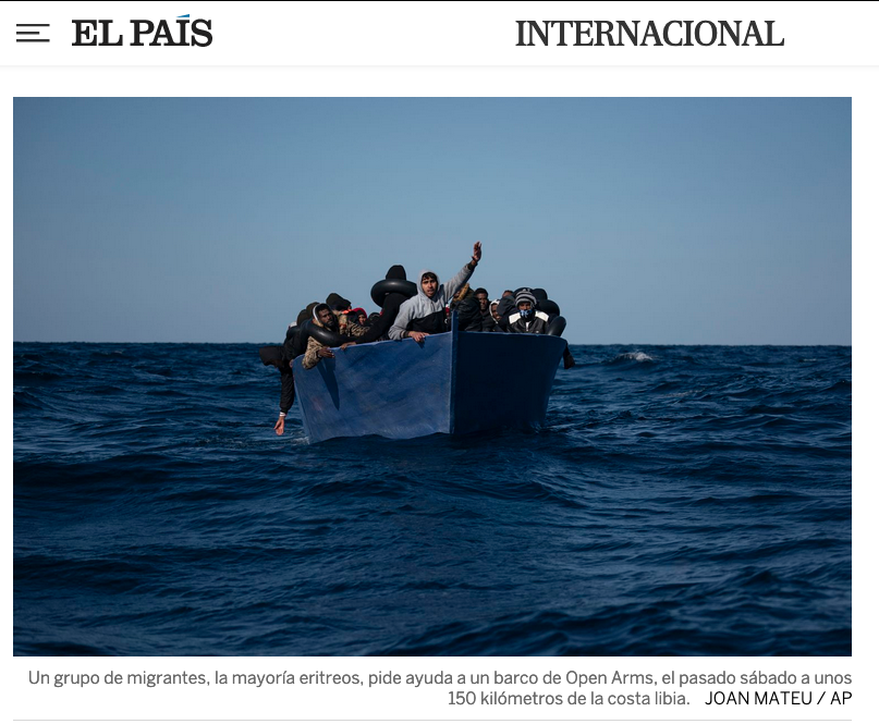 A group of Eritrean migrants on an Open Arms boat in the Libyan sea, with some hanging over the sides, and one person waving, against a blue sky and ocean background.