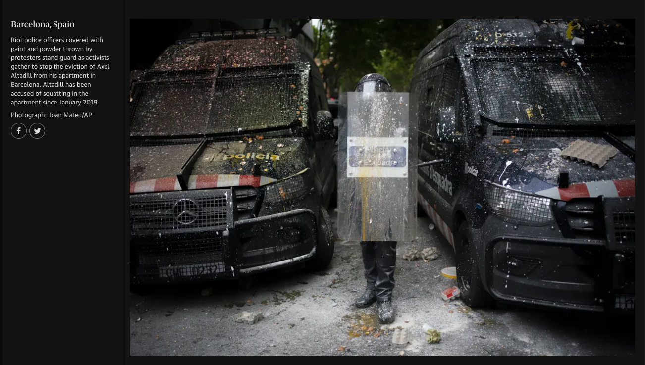 Police vehicles covered with paint and debris in an urban street, with a person holding a transparent riot shield positioned between the vehicles.