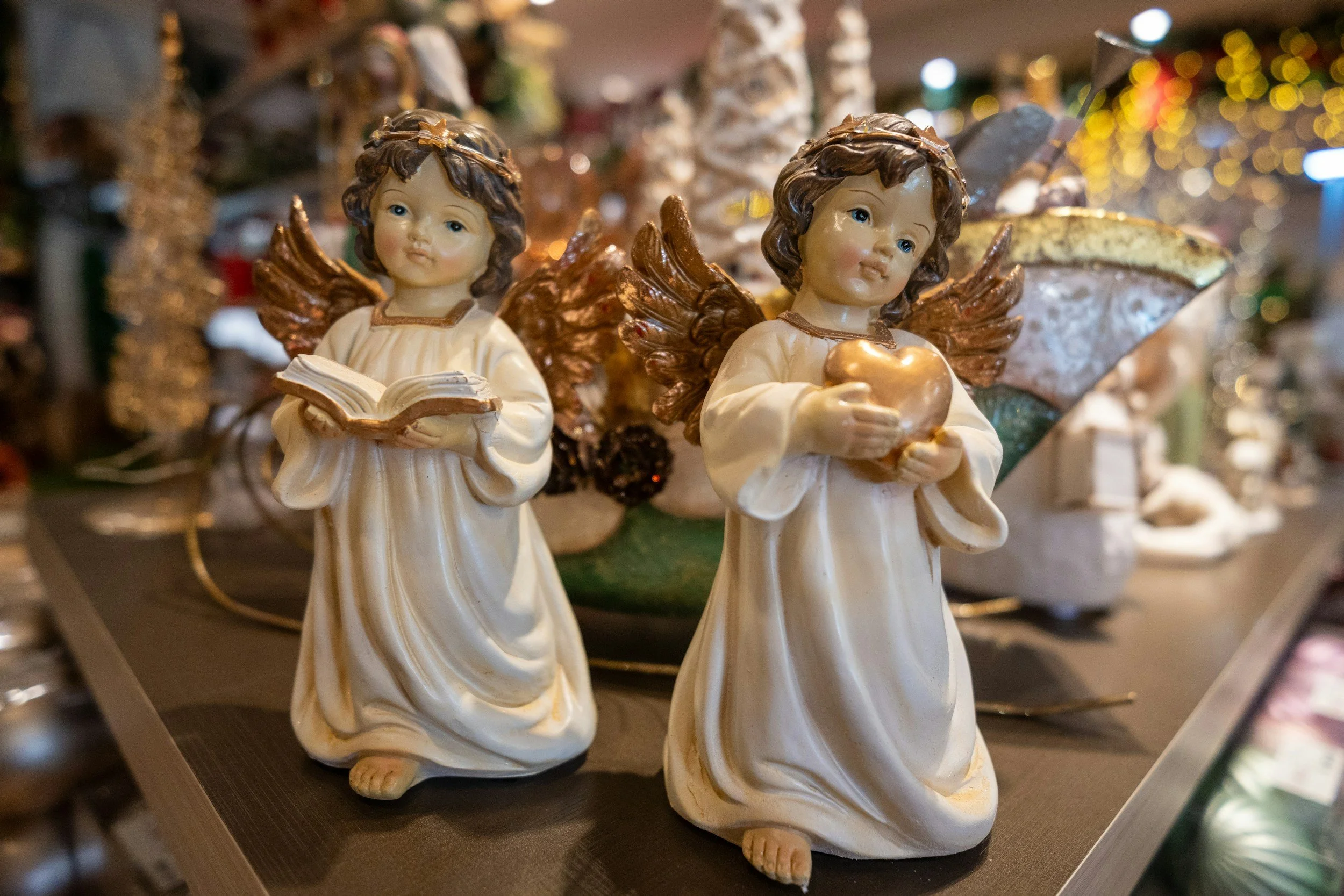 Dos figuras angelicales de cerámica, una con libro y otra con corazón, en un decorado navideño con luces y adornos.